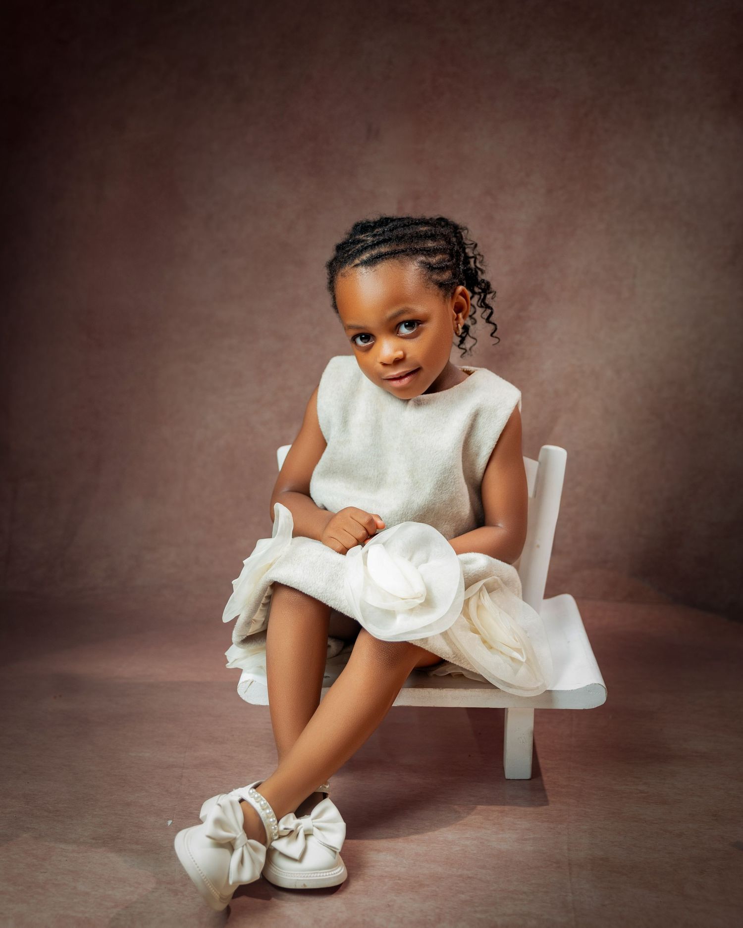 Adorable young Black girl in white sleeveless dress seated on white chair with skirt detail with a Children’s photographer in North York