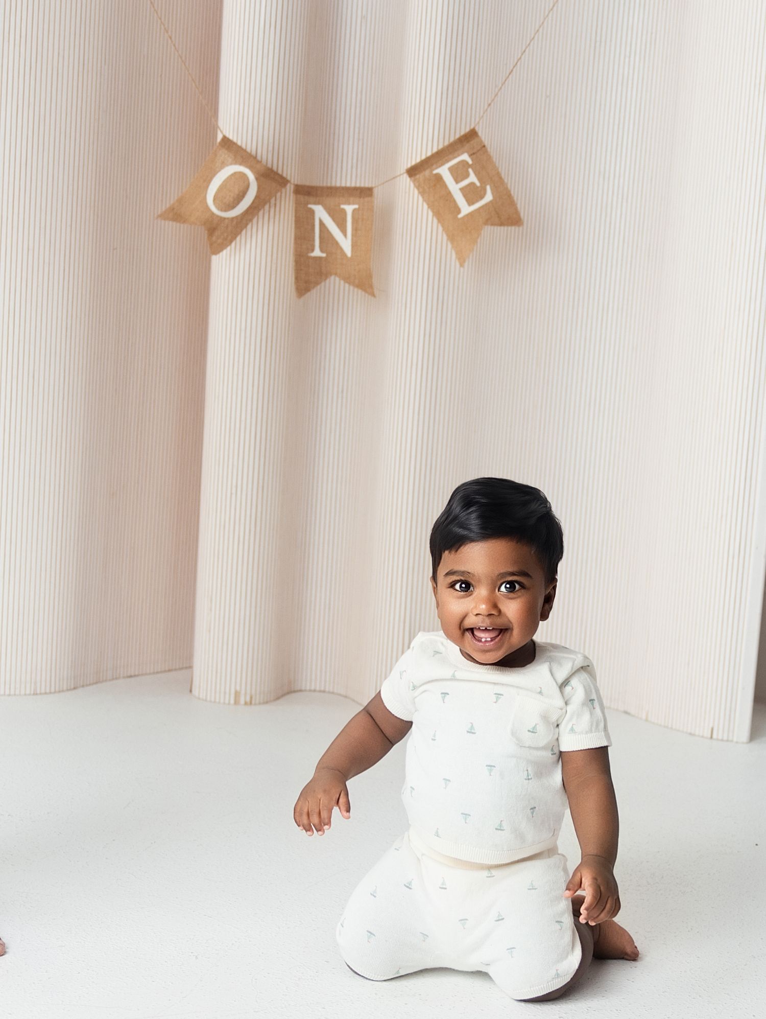 Smiling baby boy in white outfit sits before a burlap ONE banner at his first birthday photo shoot.