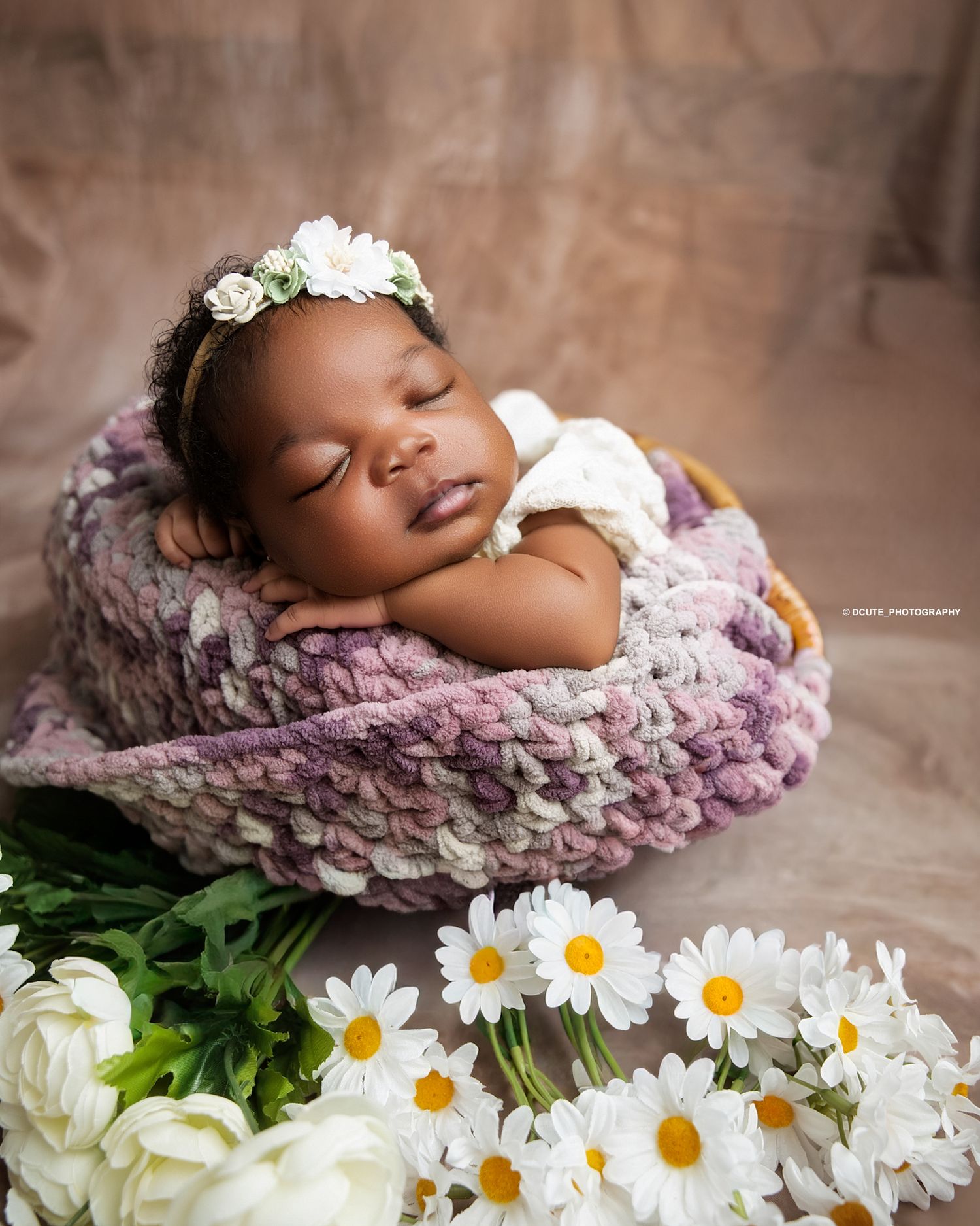 Sleeping newborn baby in a knit basket with white daisy flowers and floral headband in a studio portrait.
