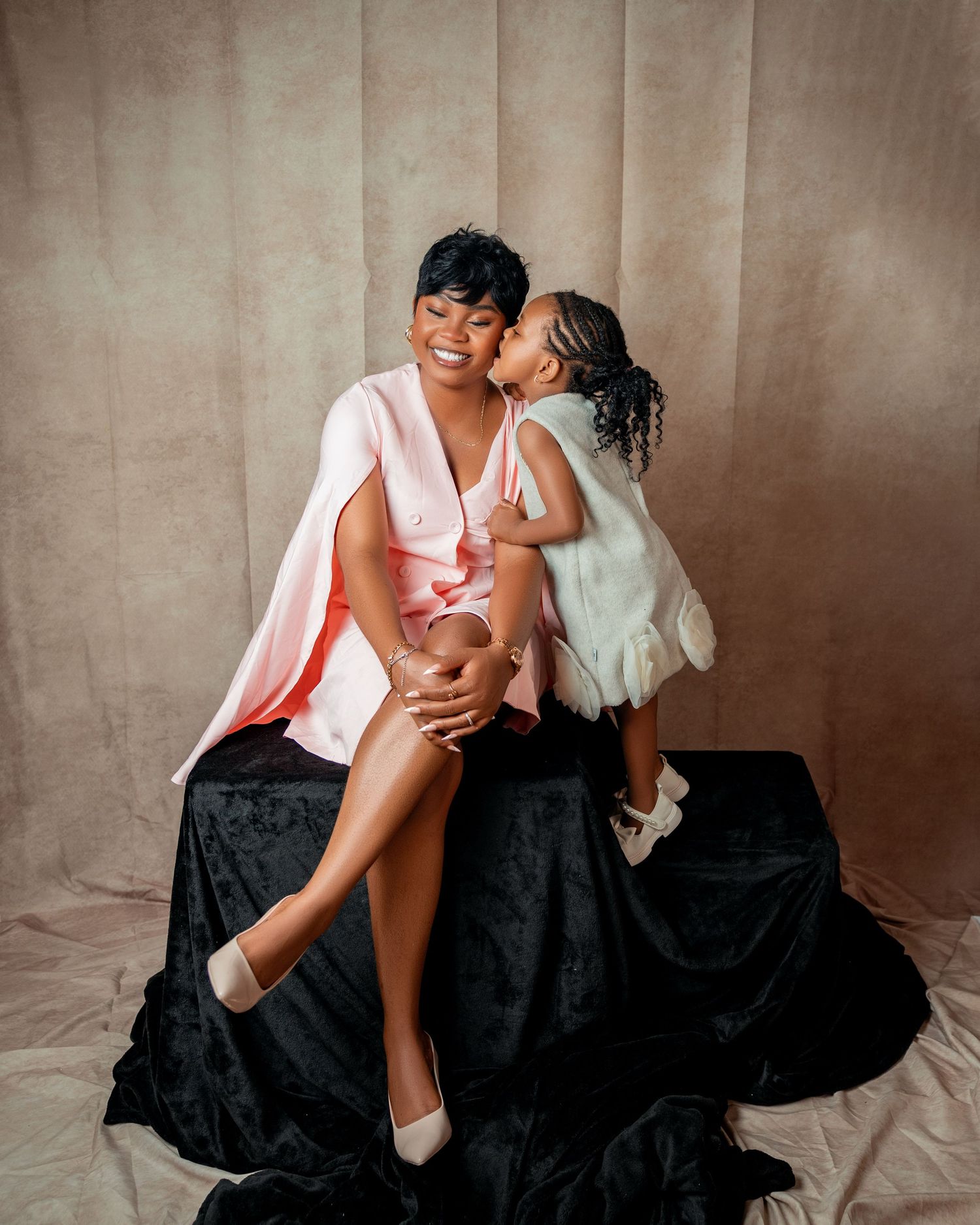 Smiling woman in pink outfit sits on black velvet surface while young girl in white dress kisses her cheek in toronto birthday photography
