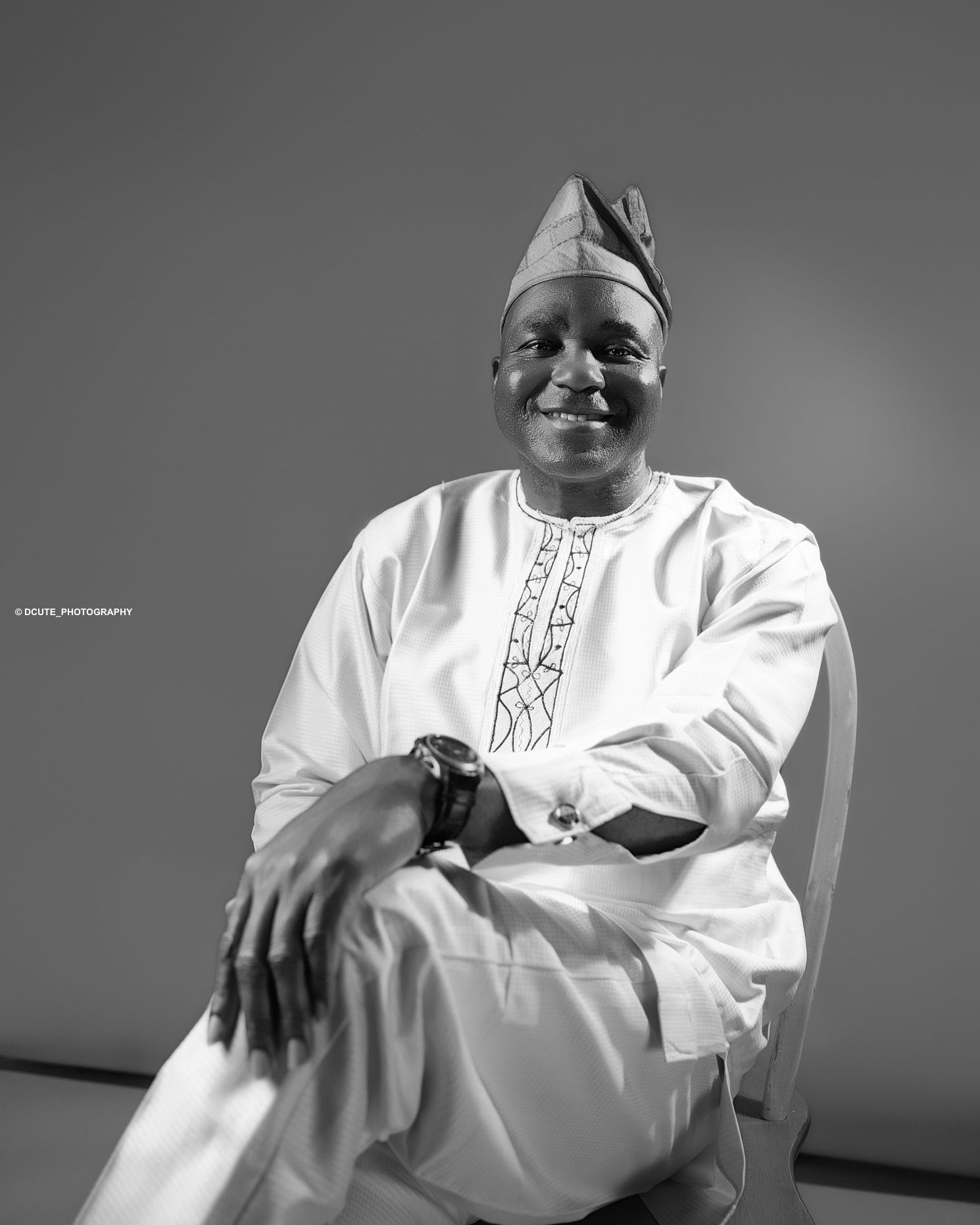 Smiling elderly man in traditional white Nigerian agbada outfit seated, captured in black and white portrait of toronto photoshoot price