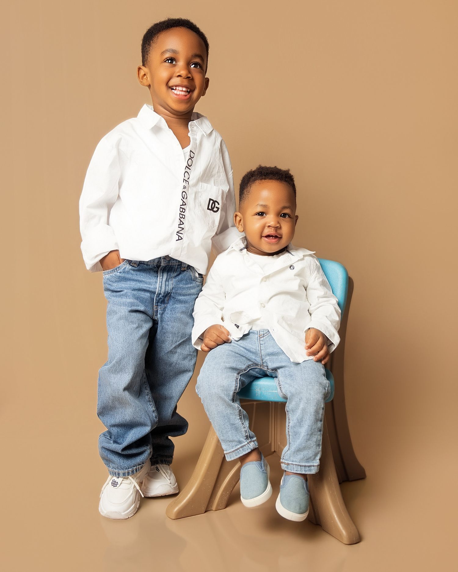 Two young boys in matching white Dolce & Gabbana shirts and jeans pose for a studio portrait photo.