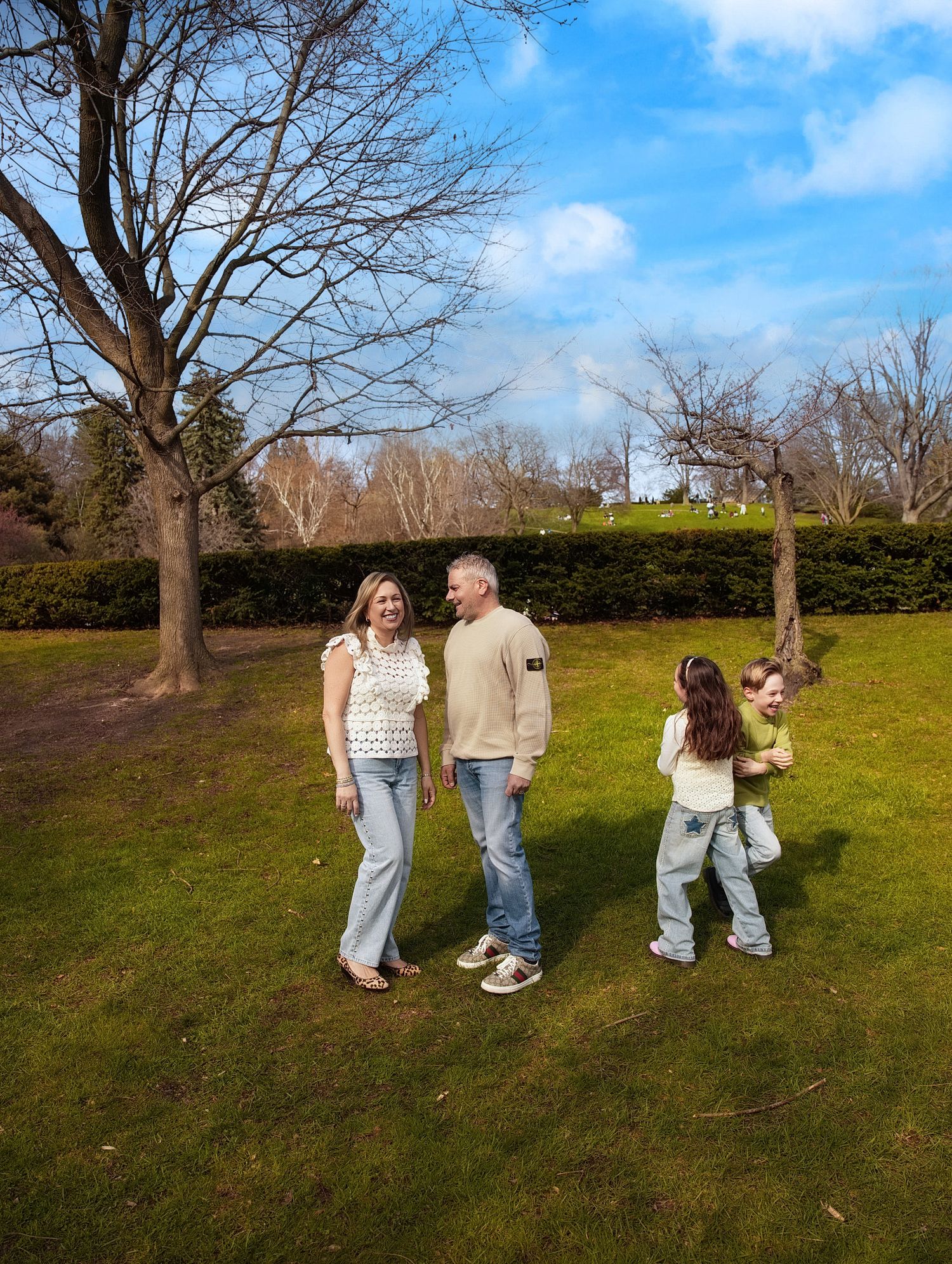 Happy family photoshoot in Toronto, Family of four enjoying a sunny day walk on green grass in a park with bare trees.