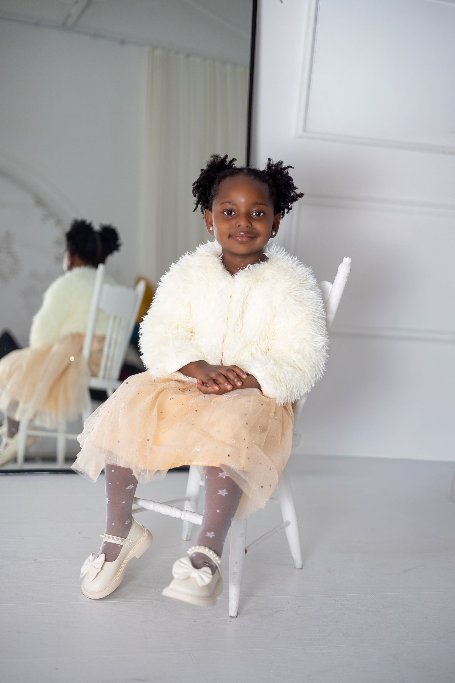 Young girl in fluffy jacket and beige tutu skirt sitting on a white chair in a bright studio captured by a North York child Photographer