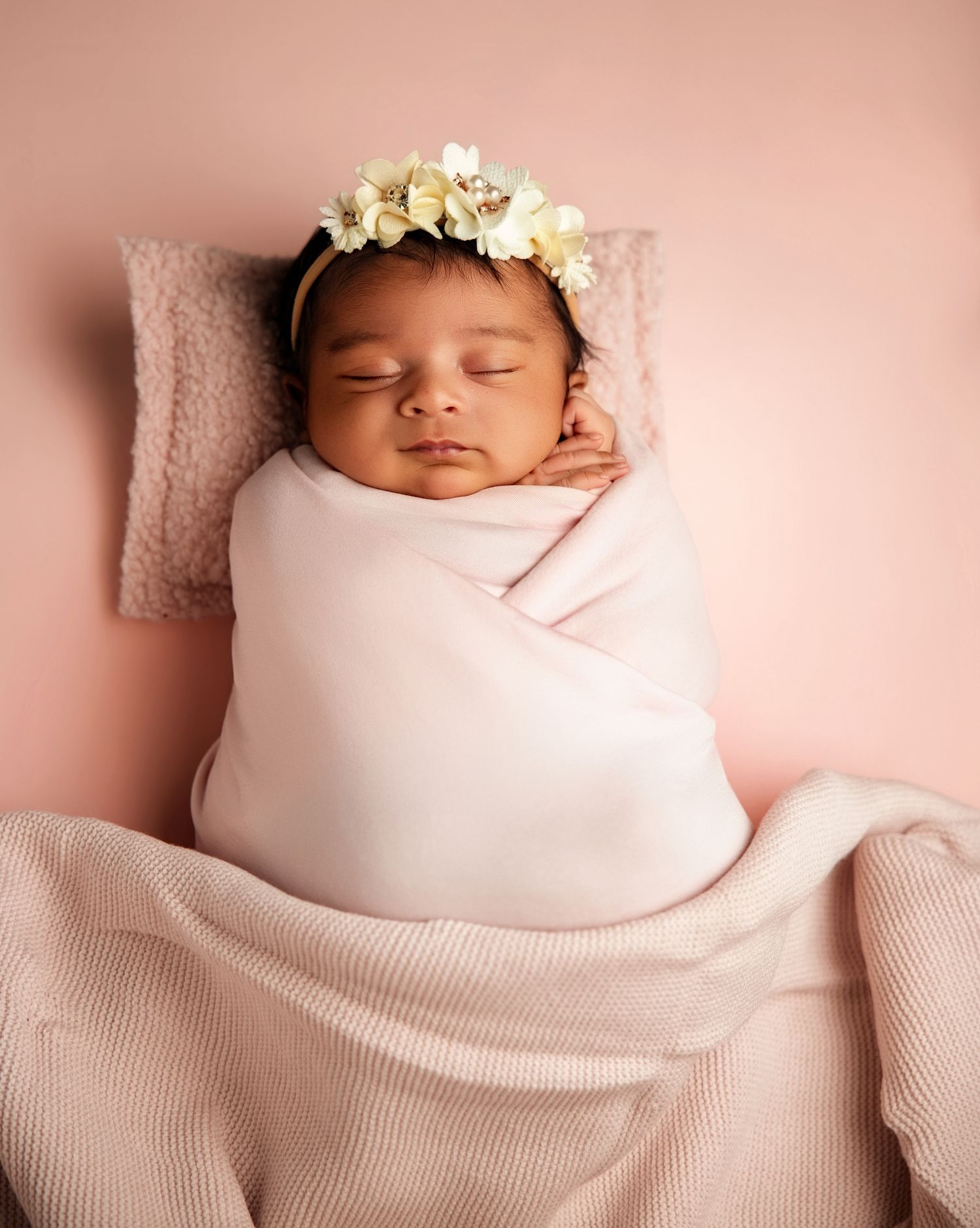 Newborn baby girl swaddled in pink blanket with floral crown sleeping peacefully on pink pillow backdrop.