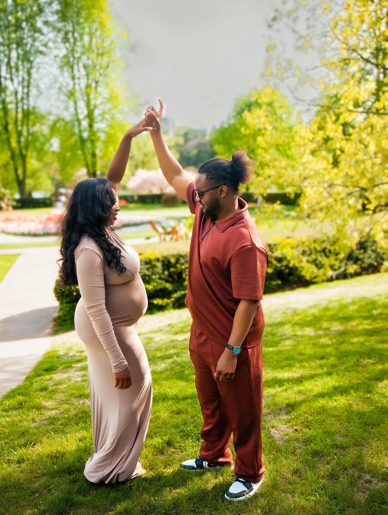 Couple dancing outdoors in sunny park, pregnant woman in beige gown with partner in rust outfit.