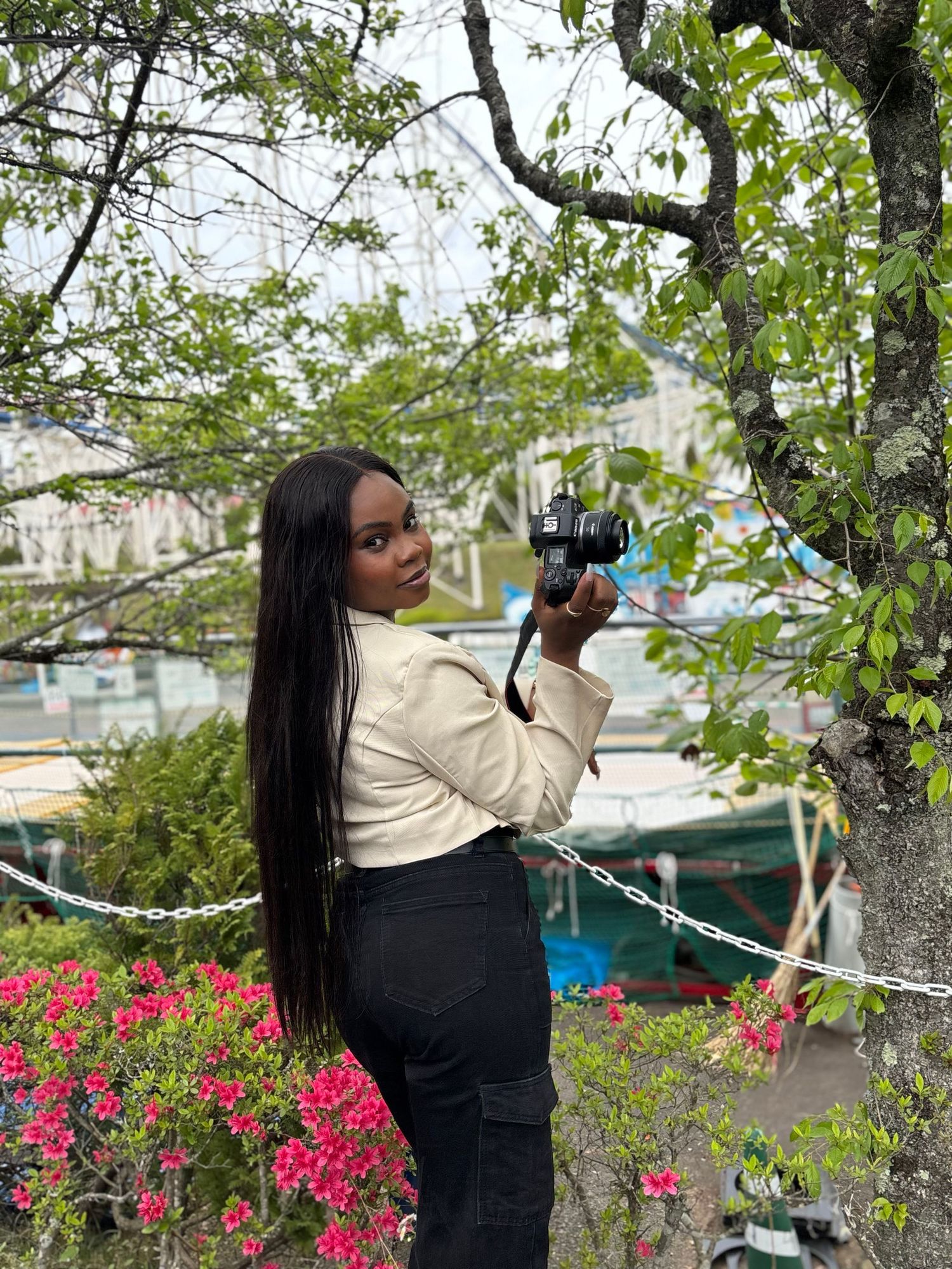 Young Black woman holds camera while posing near flowering bushes at a marina. She captures great family photos in toronto