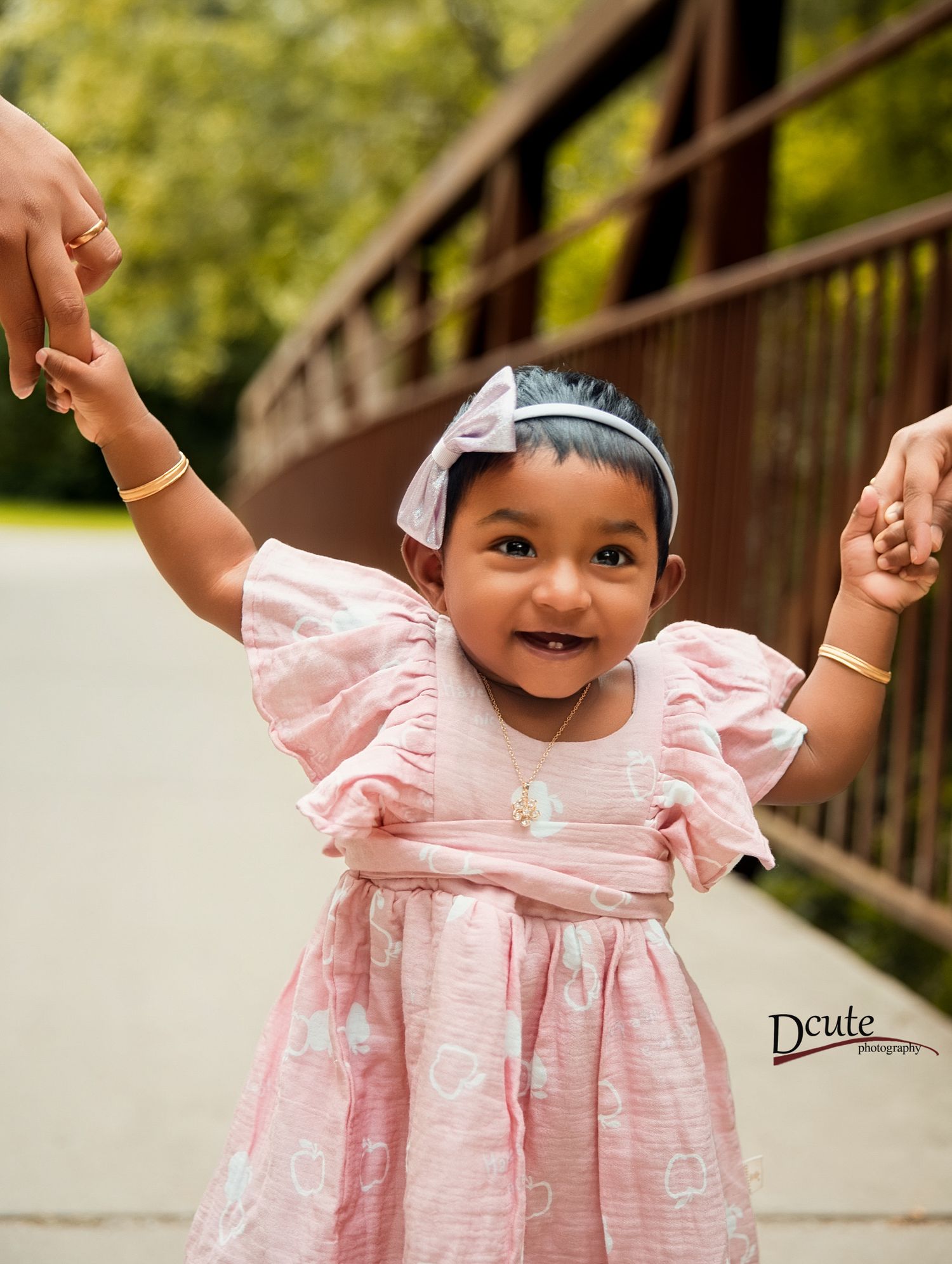 Smiling toddler girl in pink ruffled dress with bow headband holding parents hands outdoor family photoshoot in toronto