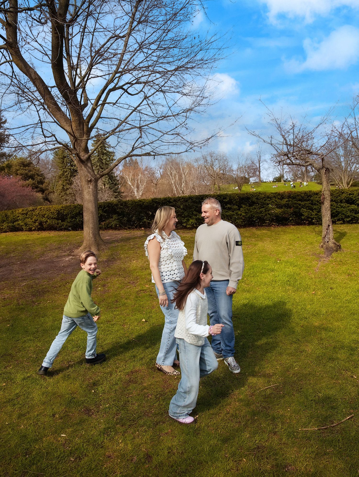 White family of four walking on green grass in park with bare trees and blue sky in background.