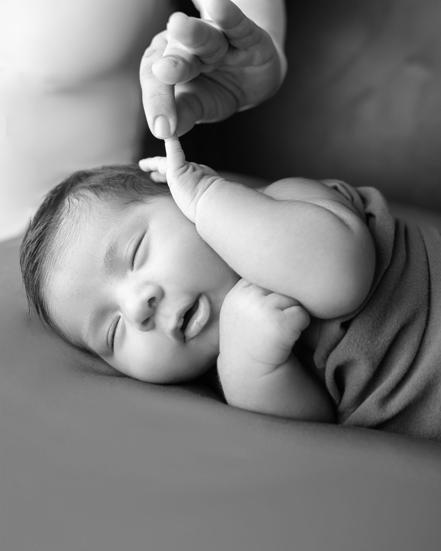 Black and white close-up of sleeping newborn gripping adult finger gently.