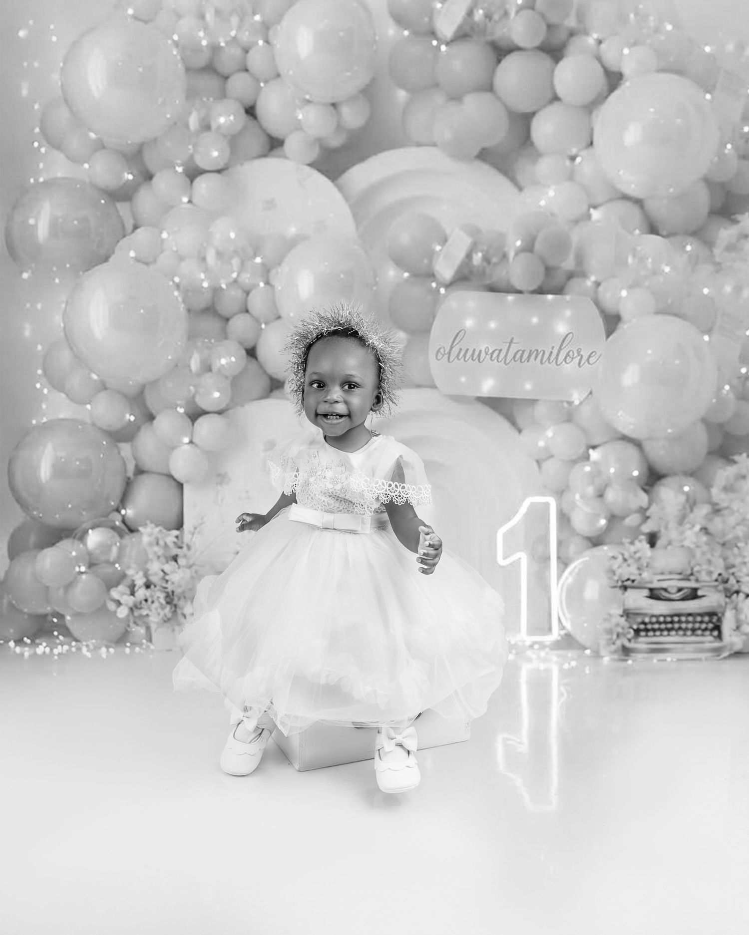 Toddler girl in white dress celebrating first birthday with balloon backdrop in black and white.