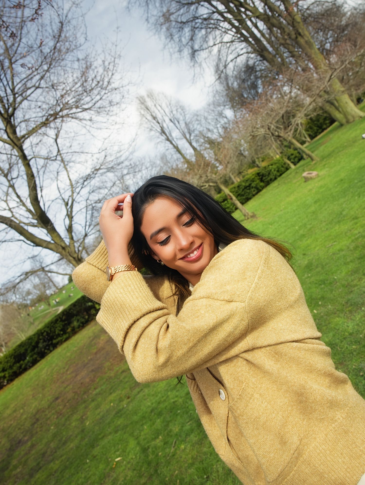 Smiling young woman in oversized yellow knit sweater posing playfully in a lush green park on a sunny day in relaxed outdoor birthday photos