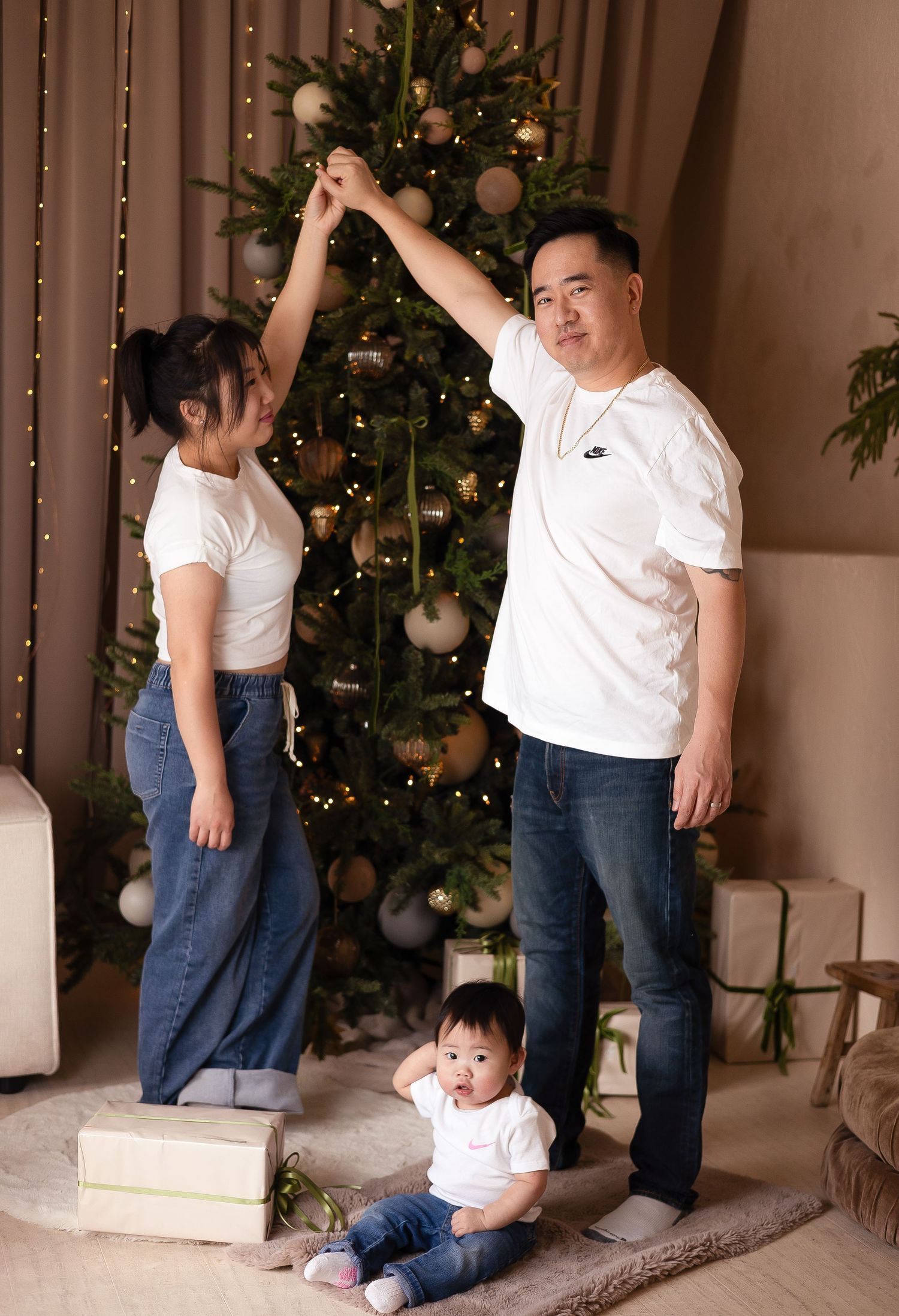 Asian couple dances holding hands above their baby seated near a decorated Christmas Family photography near Downtown Toronto