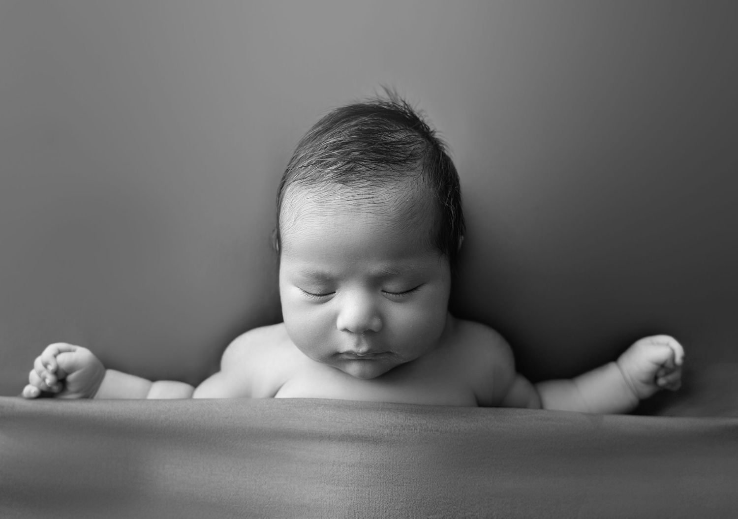 Black and white newborn portrait of a sleeping baby with dark hair, arms spread wide, peaceful expression. IN A WEST TORONTO PHOTOGRAPHER PO