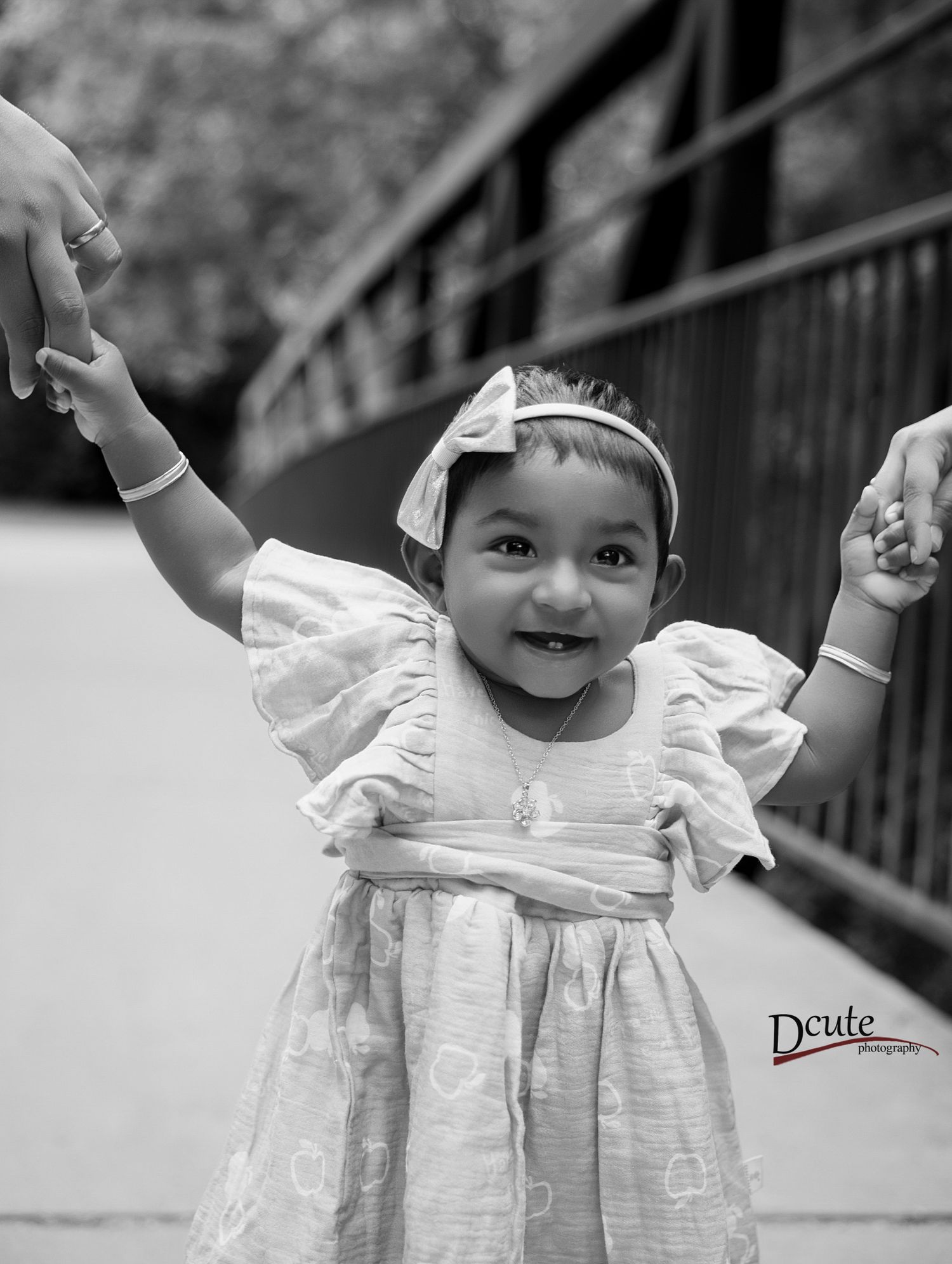 Black and white photo of a happy baby girl in a white dress holding adults hands outdoors by a kids photography in North York
