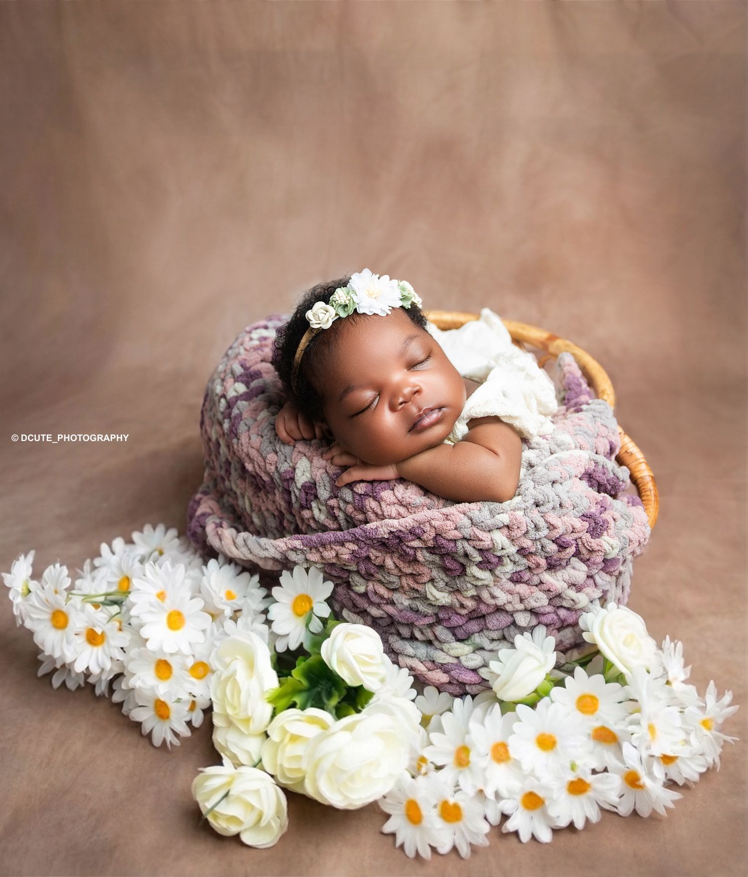 Sleeping newborn baby in a knitted basket surrounded by white daisies and roses, wearing a floral headband.