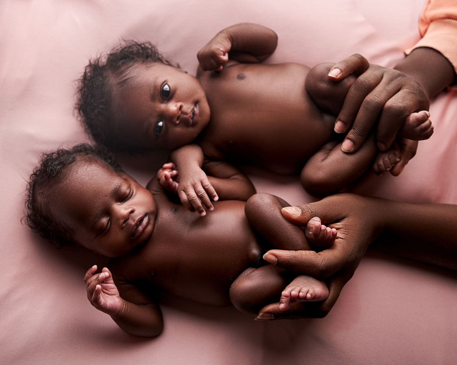 Twin newborn babies lying on pink fabric, held gently by an adult hand in a tender portrait in a Toronto newborn photography