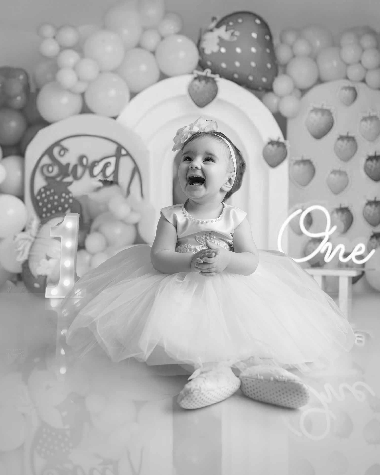 Black and white photo of baby girl in tutu dress laughing at first birthday party with balloon decorations.