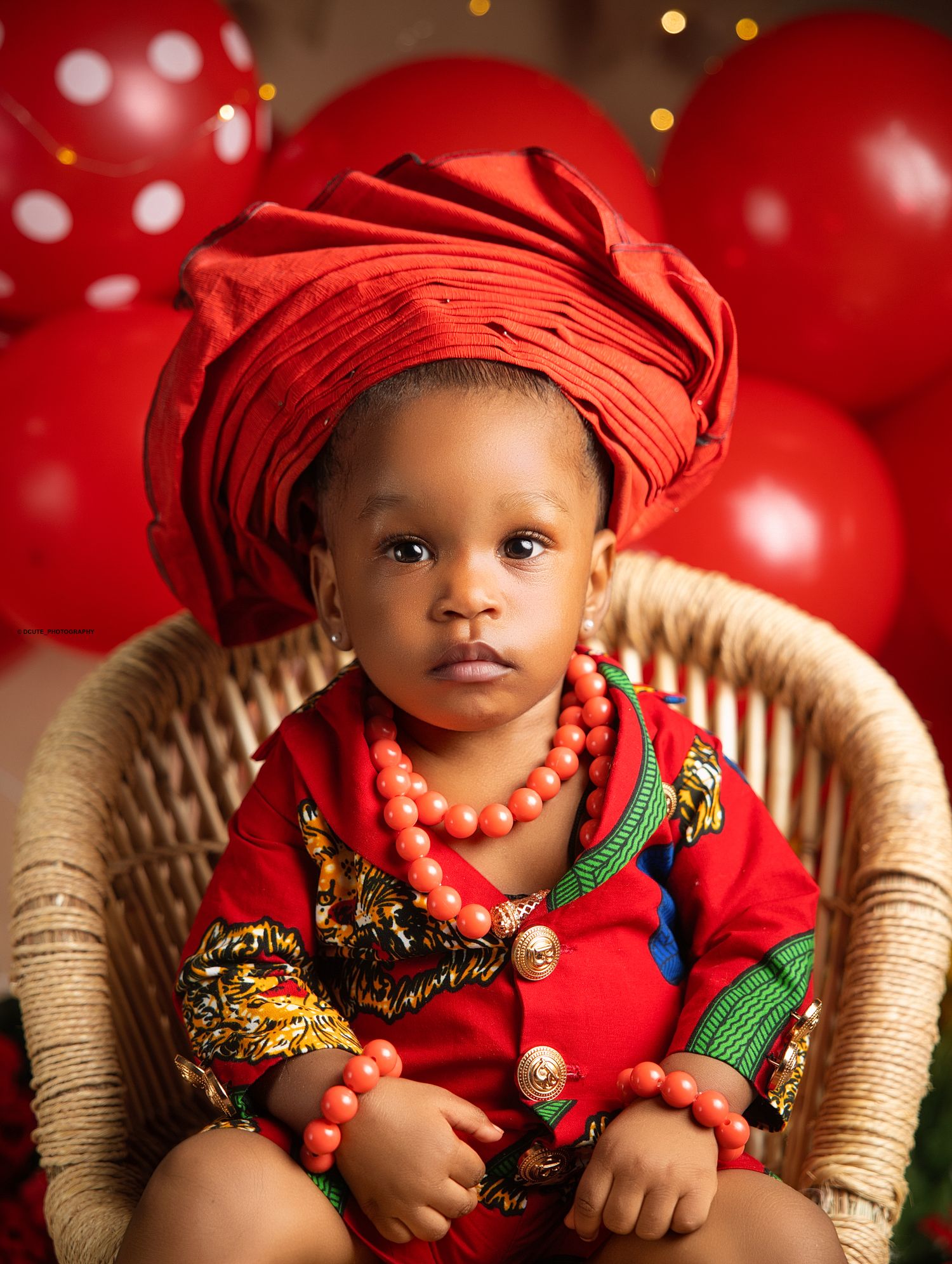 Baby dressed in traditional red African attire with coral beads sits in chair with red balloons in Children’s photography Toronto session