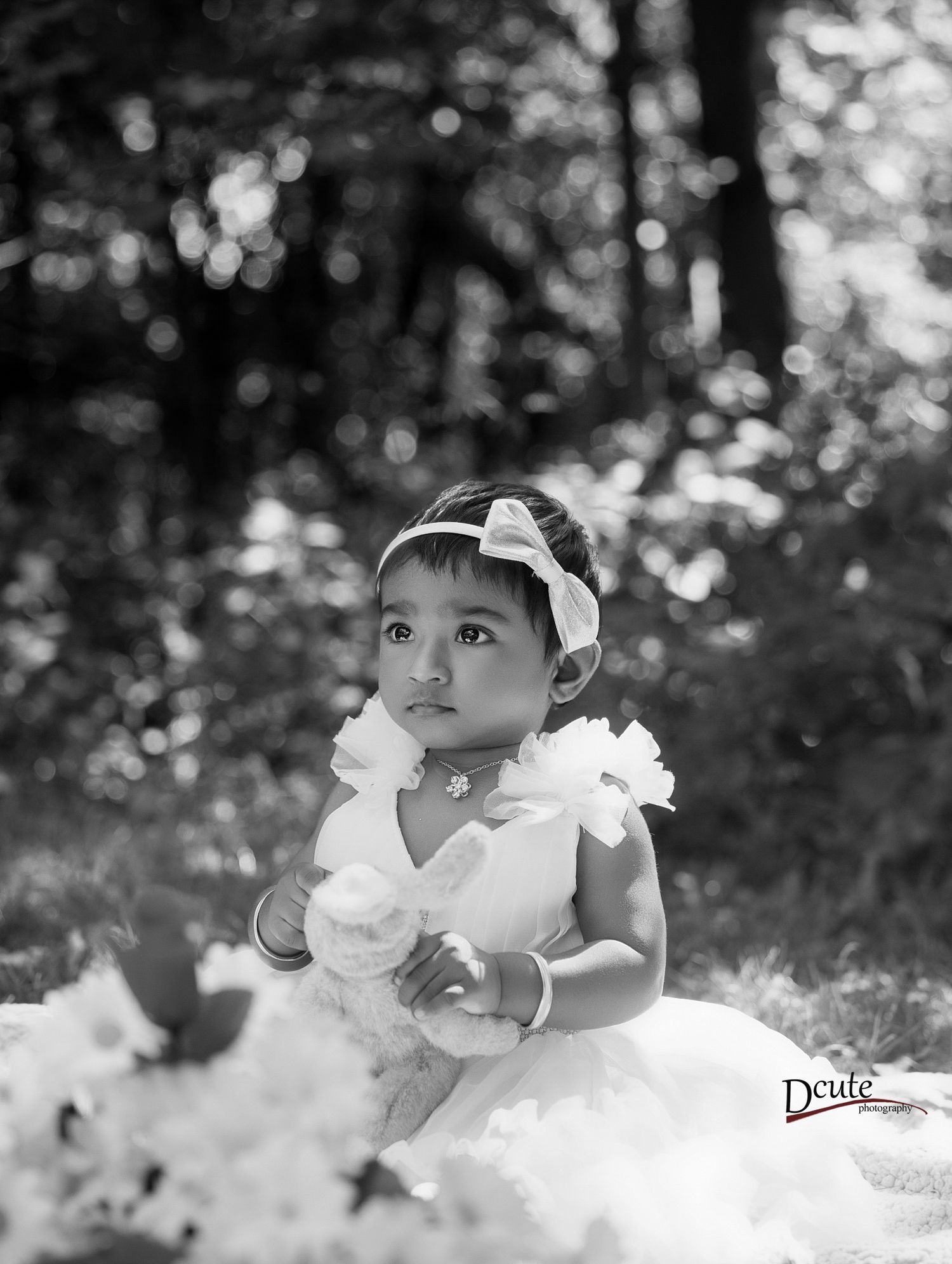 Black and white portrait of a toddler girl in a white dress and floral headband sitting outdoors among flowers.