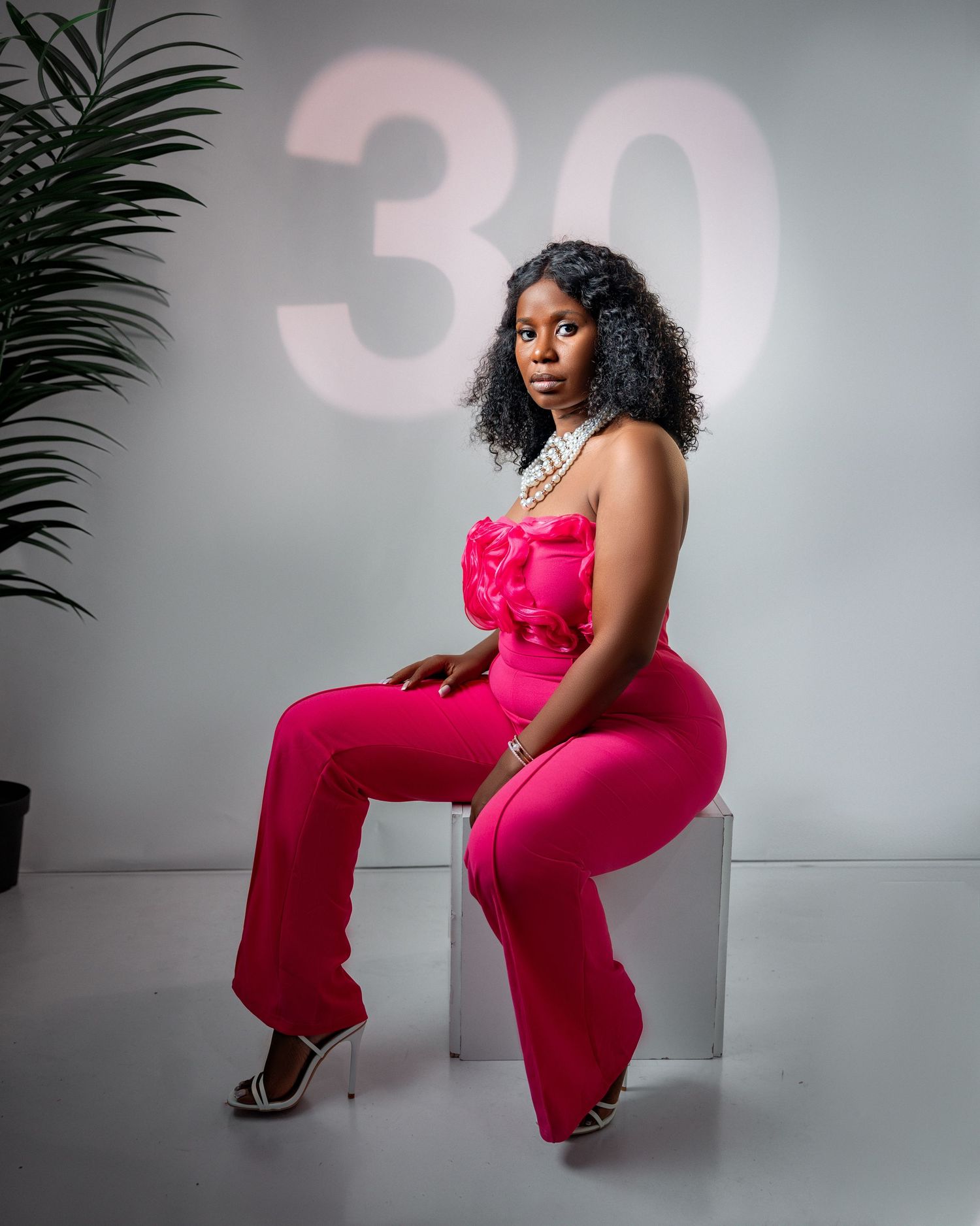 Woman in hot pink outfit seated on white cube with number 30 on wall behind a creative birthday photoshoot in Toronto studio