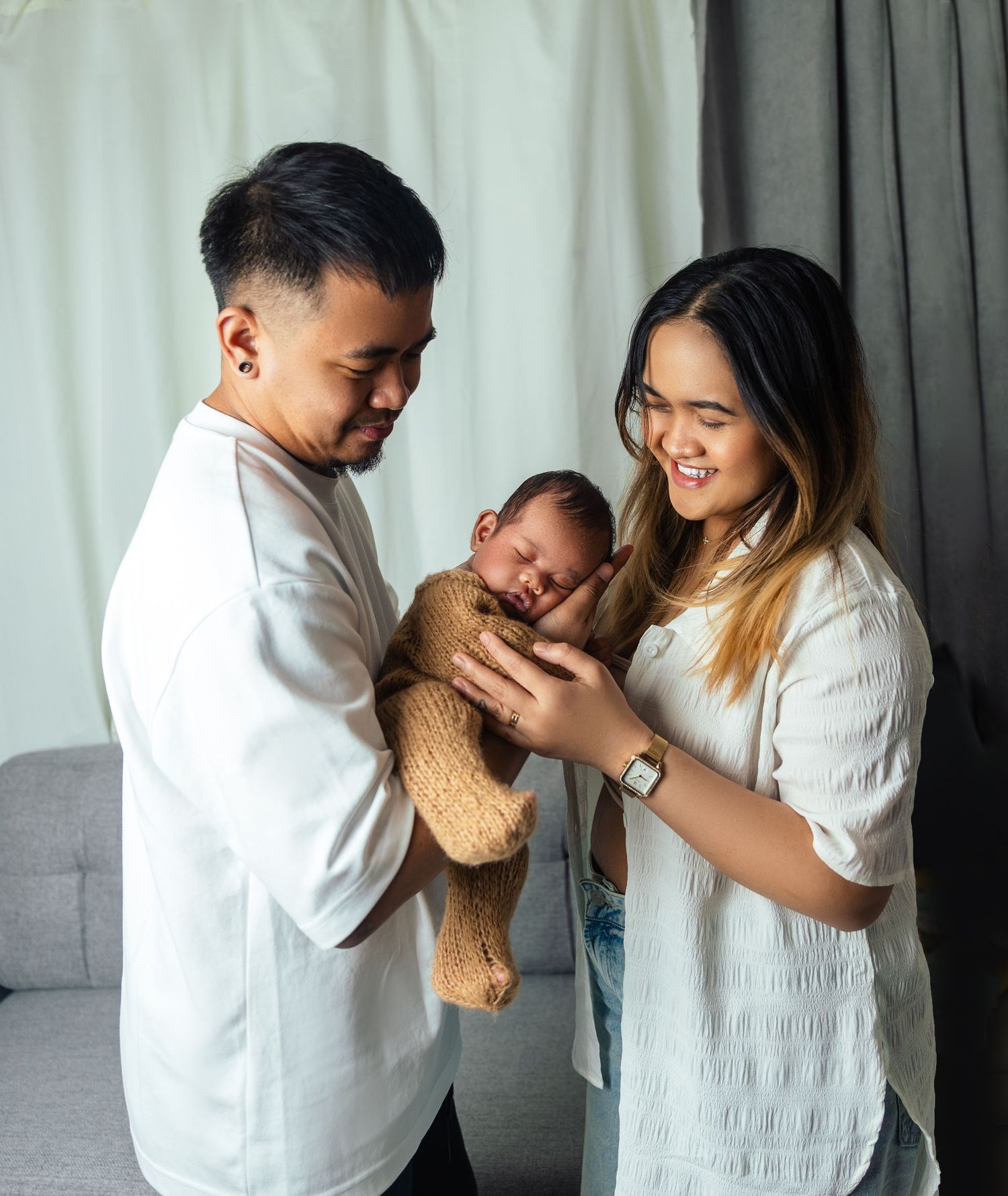 Happy parents in white outfits lovingly hold their sleeping newborn baby wrapped in a brown knit blanket.