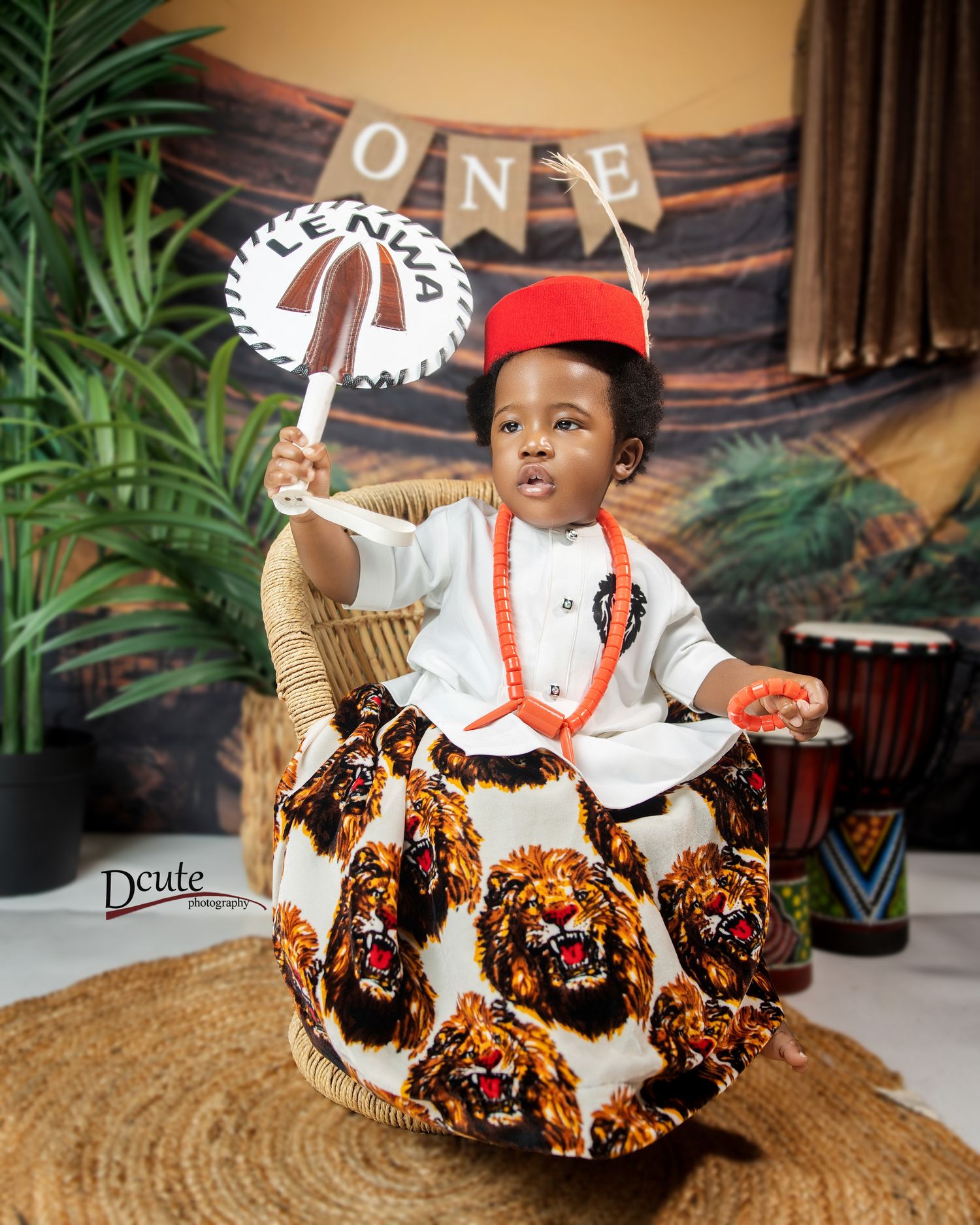 Toddler in Igbo cultural outfit holds fan and wears red cap at first birthday.