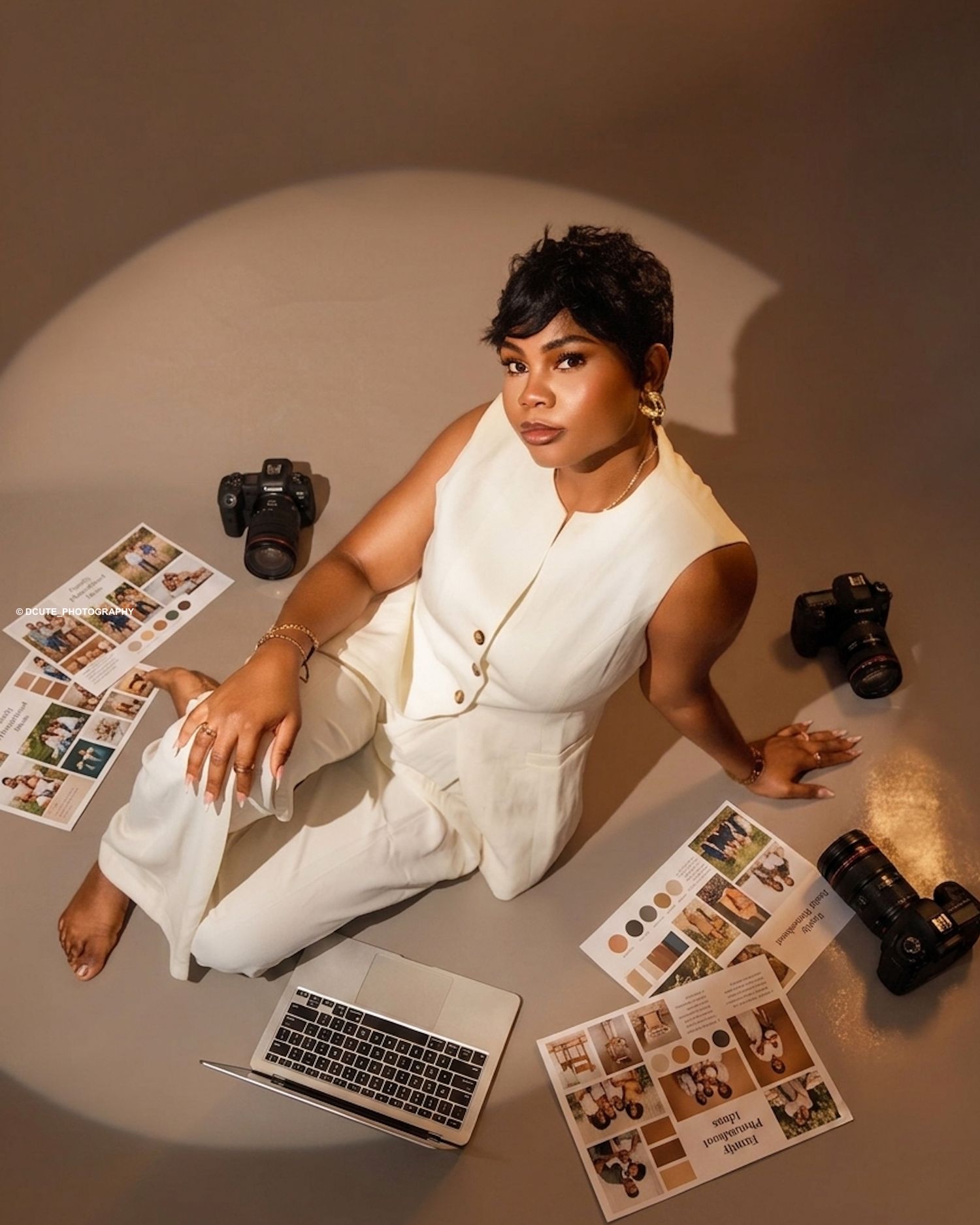 Woman in white outfit sits on floor surrounded by cameras, laptop, and photo prints in studio setting.