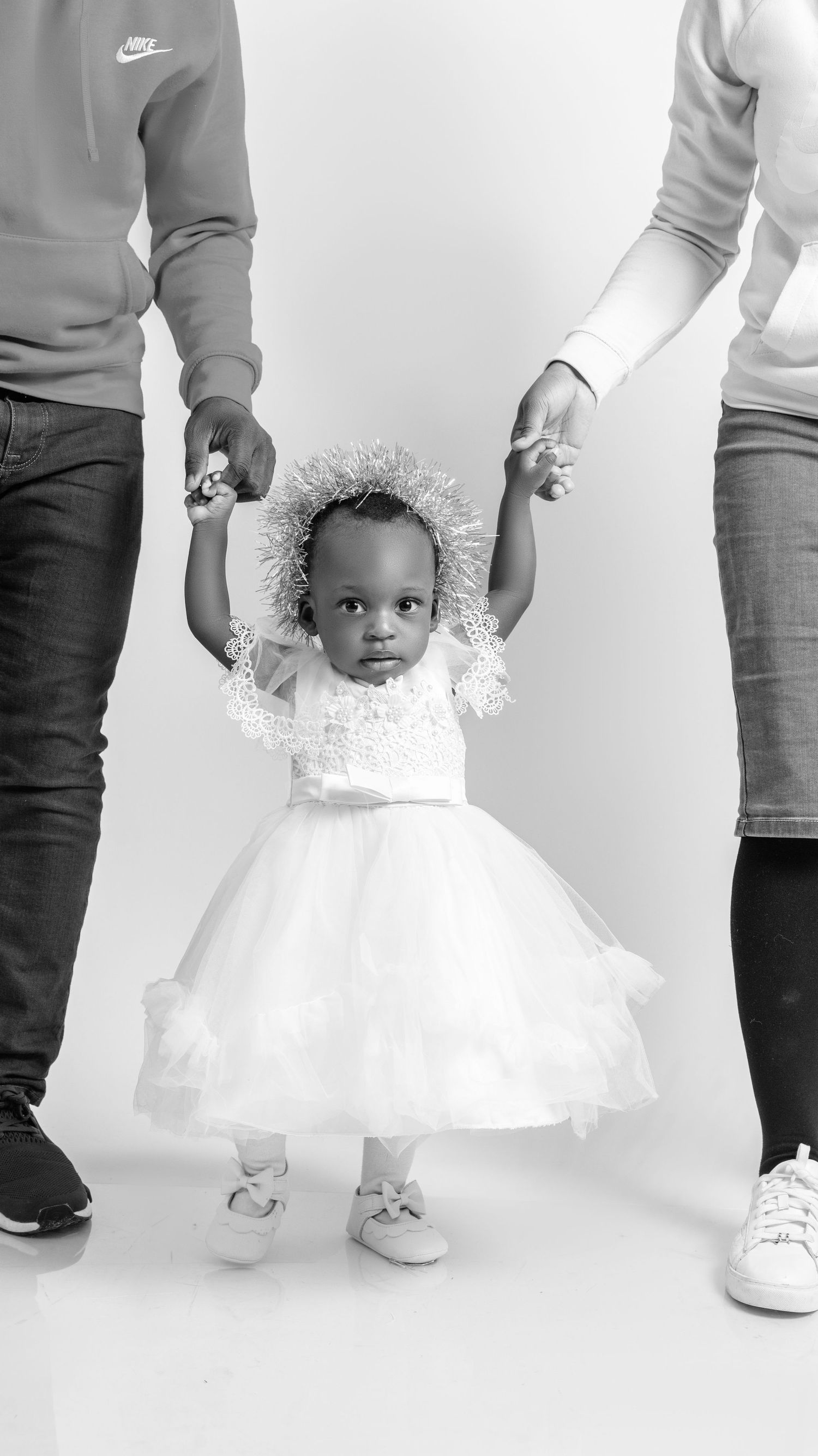 Baby girl in white tutu dress with silver crown held by two adults in black and white photo.