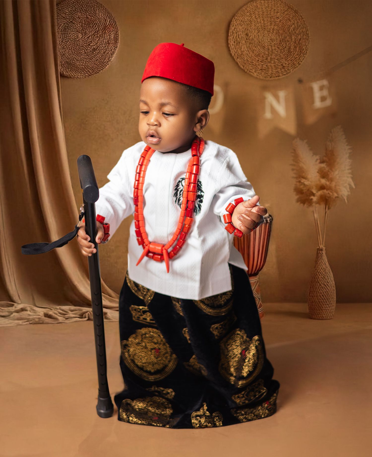 A toddler dressed in traditional Nigerian Igbo attire with red beaded necklace and red cap poses proudly.