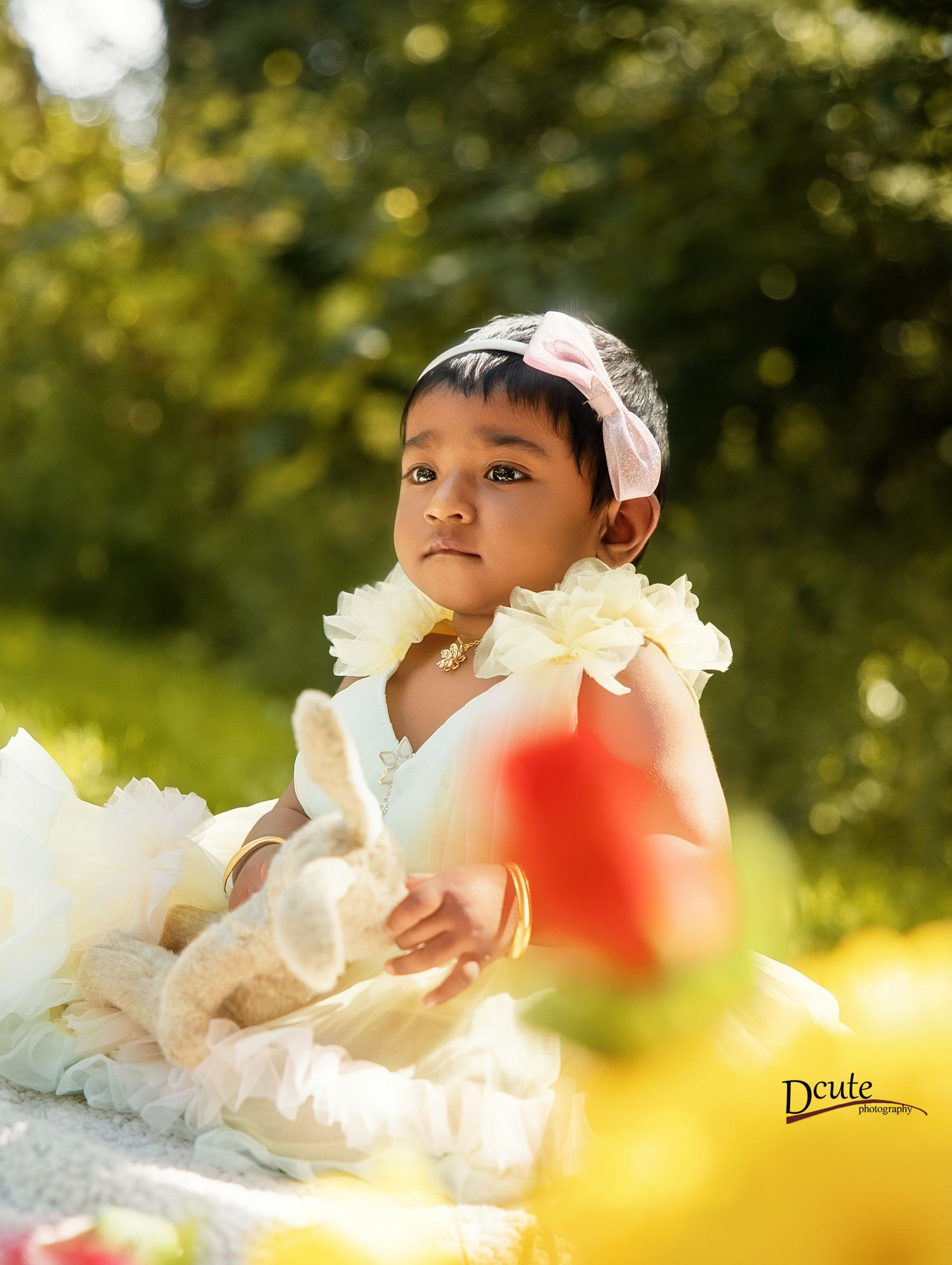 An adorable baby girl in a white ruffled dress and pink headband sits among flowers shot by a professional family photographer in Toronto