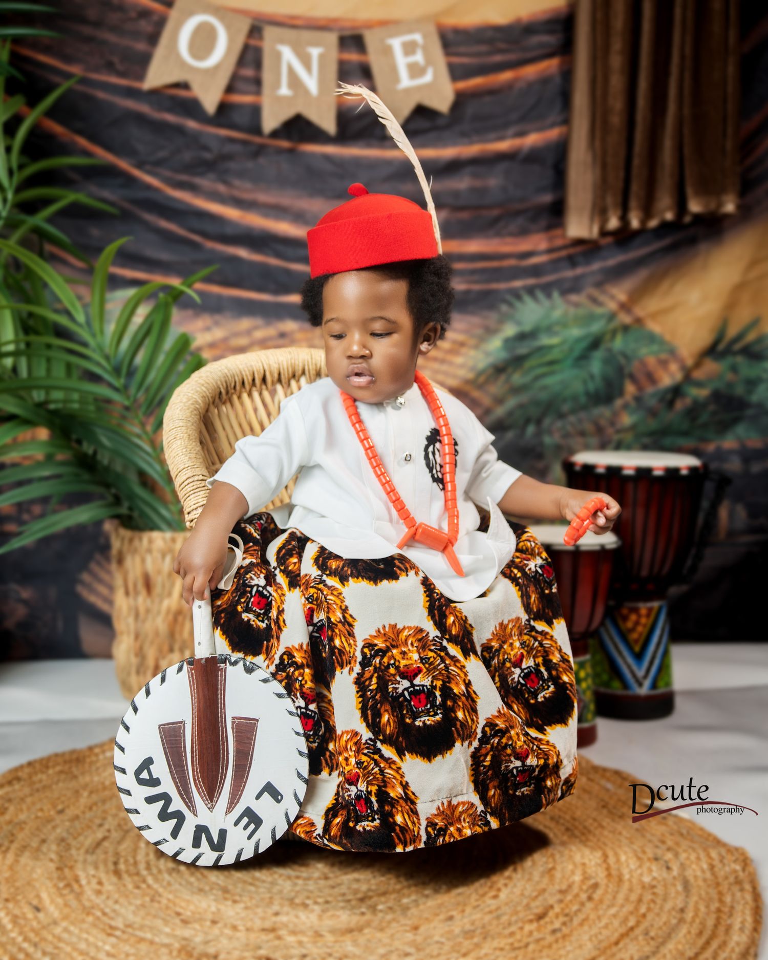 Baby boy in traditional Igbo attire with red feathered cap sitting beside a shield for first birthday in African cultural family photoshoot