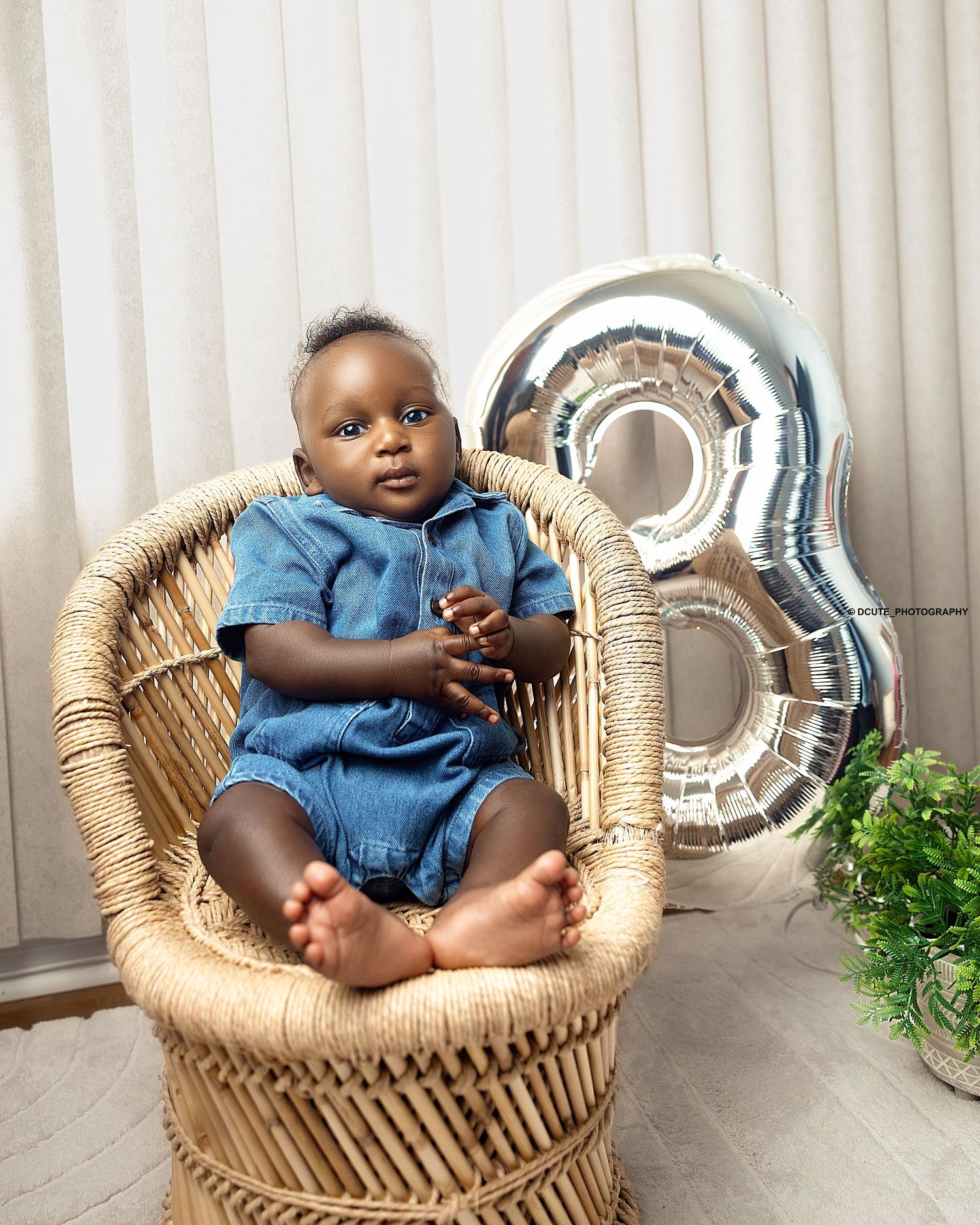 A baby in a denim romper sits in a wicker chair beside a silver number 8 balloon for a milestone photo. IN A WEST TORONTO PHOTOGRAPHER PORTF