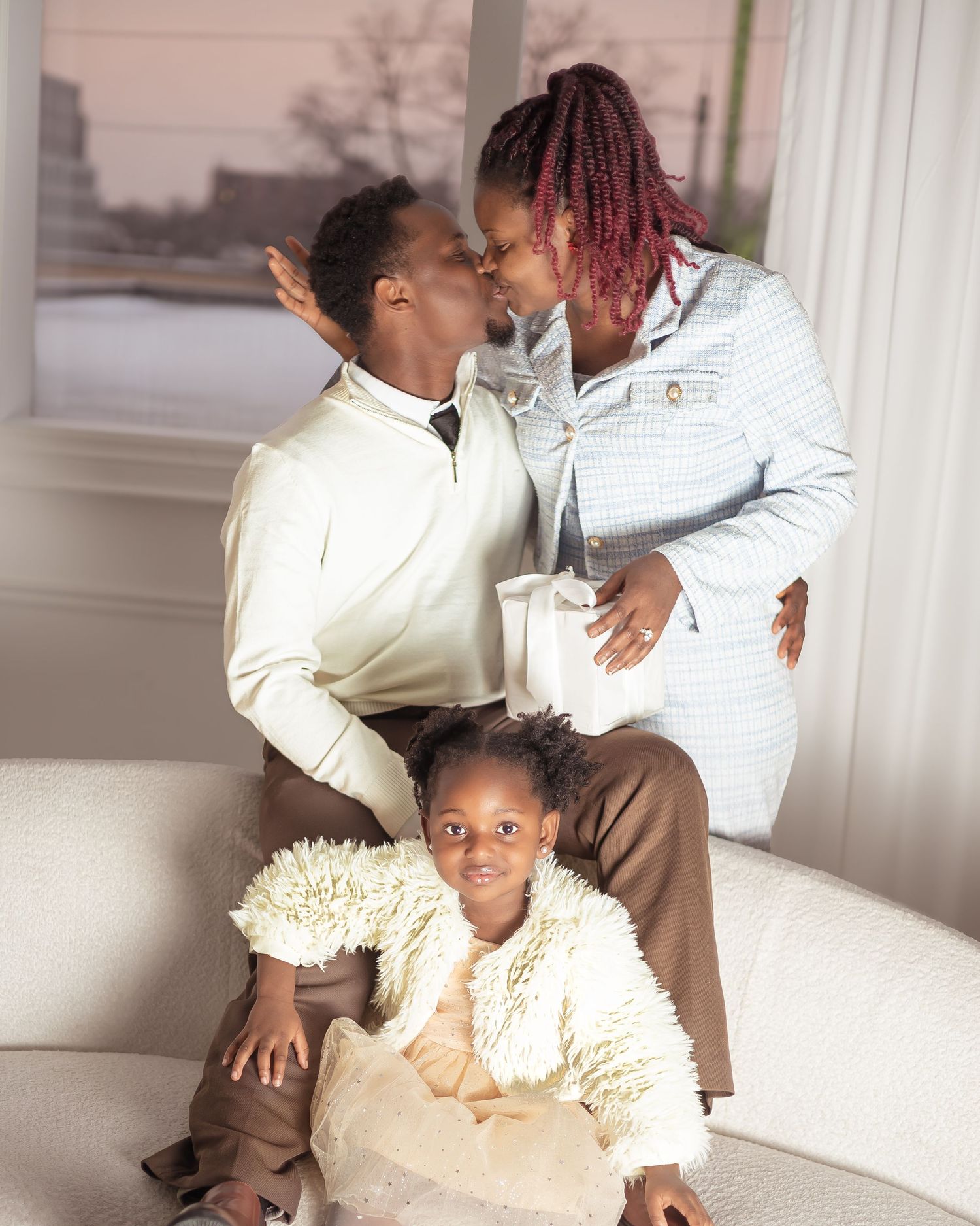 Pregnant Black woman kissing partner on white sofa with young daughter in fluffy jacket during family photo session in downtown toronto
