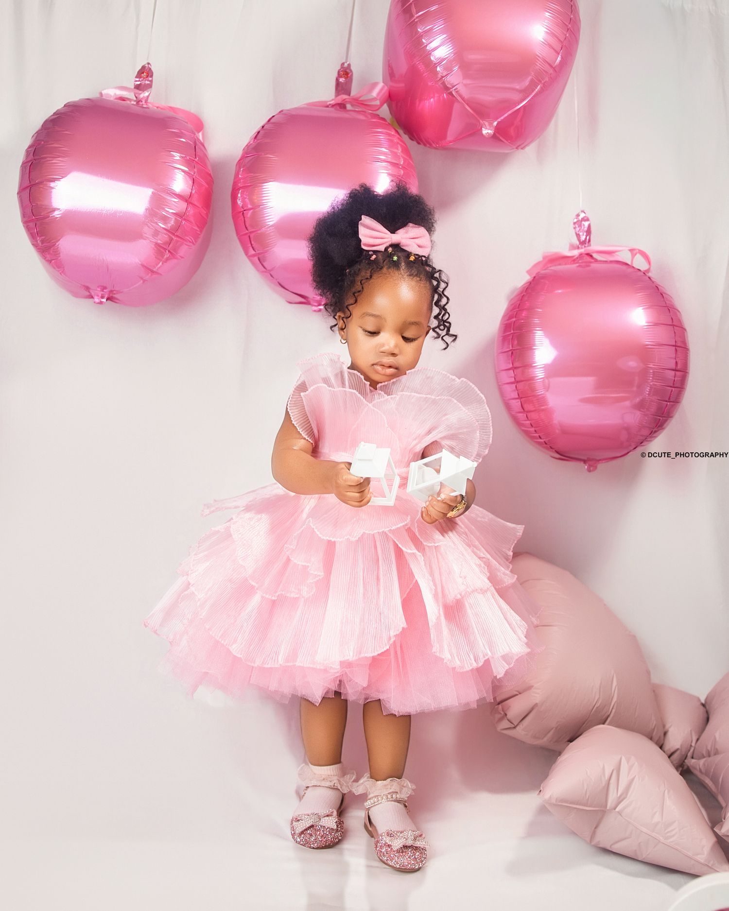 A toddler in a pink layered tulle dress holds a bow near pink balloons in a soft studio setting.