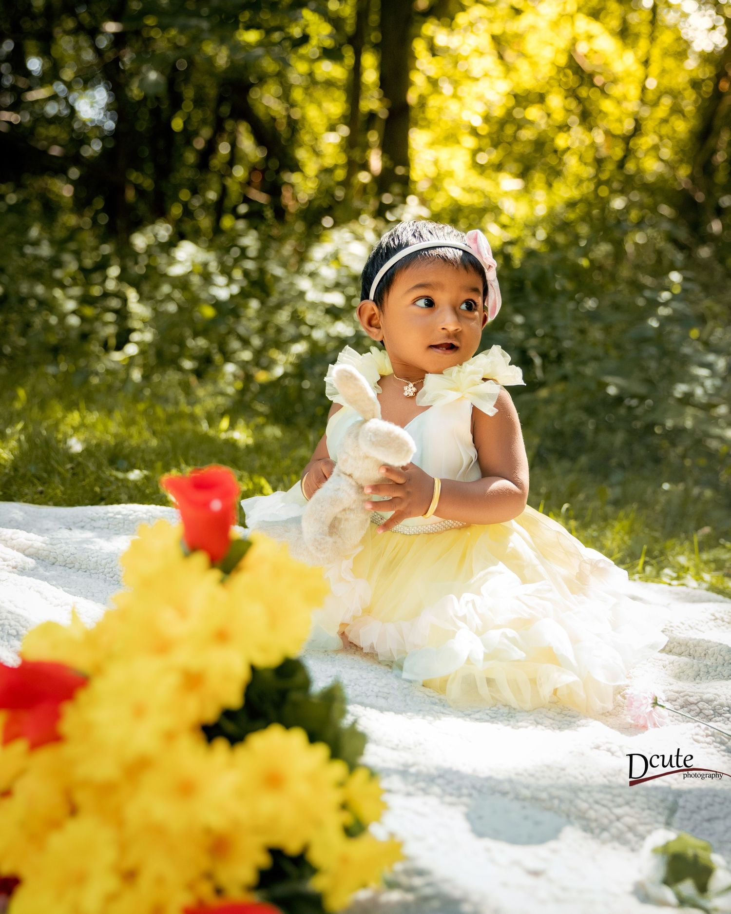 Baby girl in yellow tulle dress holds stuffed bunny among colorful garden flowers.