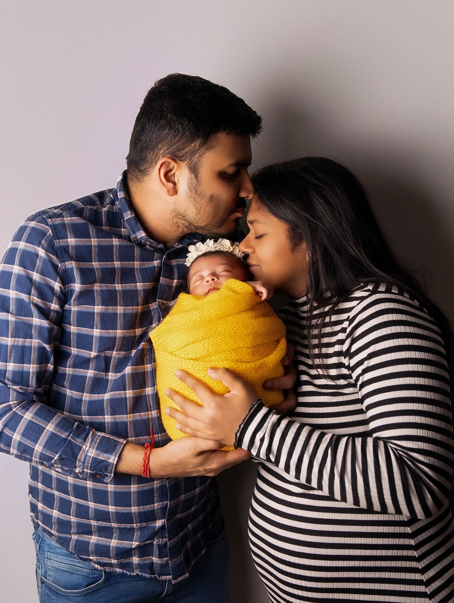 New parents tenderly kissing their newborn baby wrapped in yellow, capturing a precious family moment.