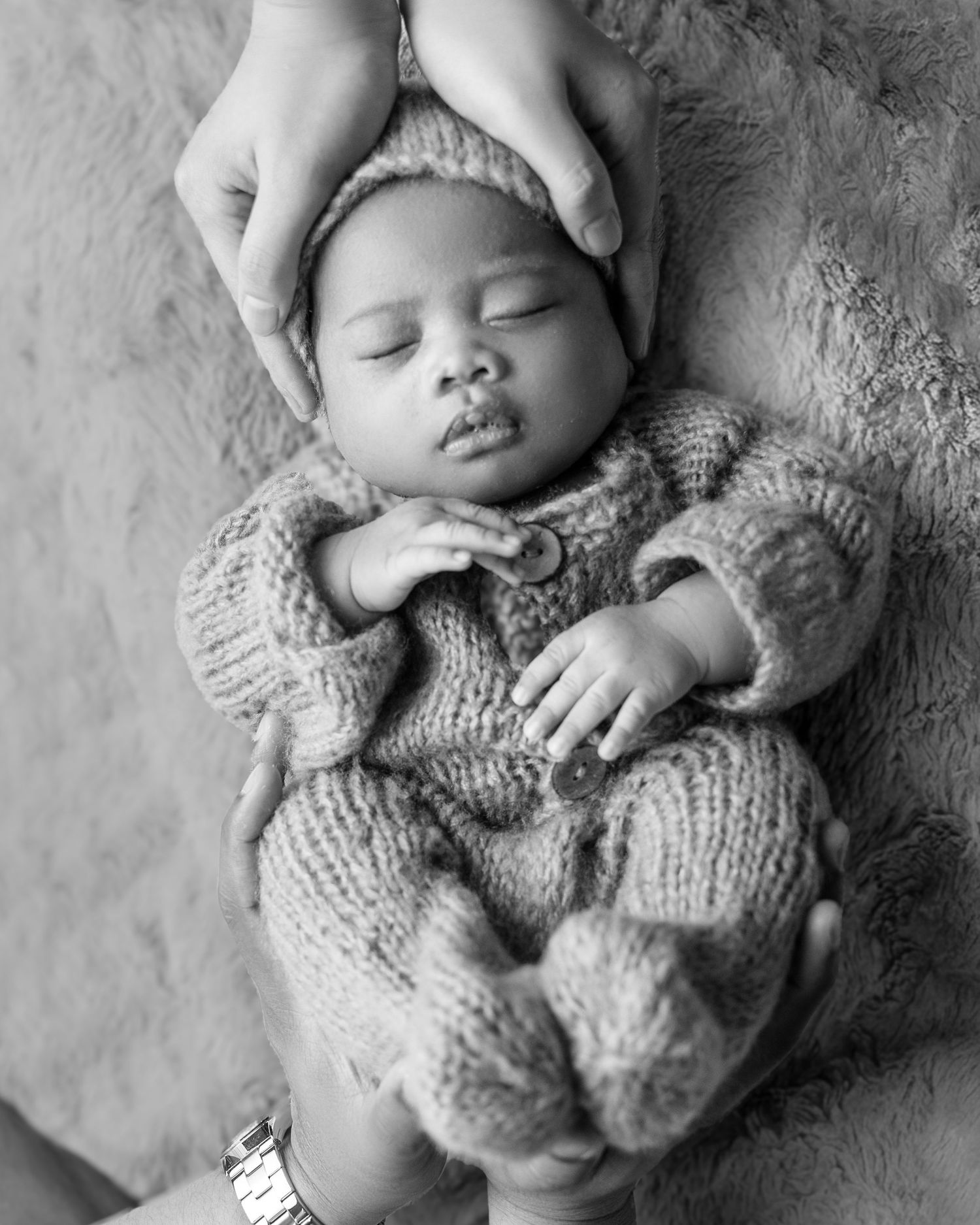 Black and white photo of sleeping newborn in knit outfit cradled by adult hands.
