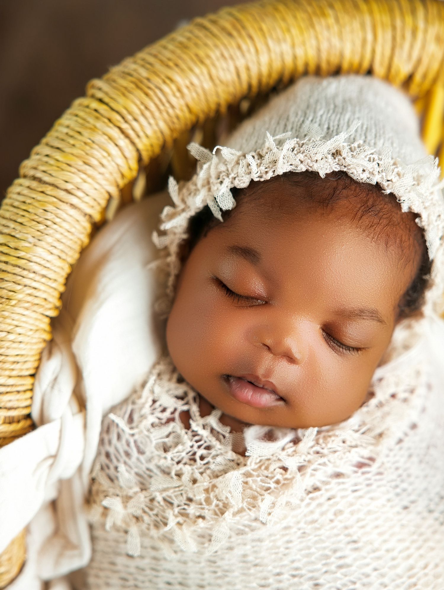 Sleeping newborn baby wrapped in cream lace swaddle wearing a delicate bonnet, nestled in a wicker basket.