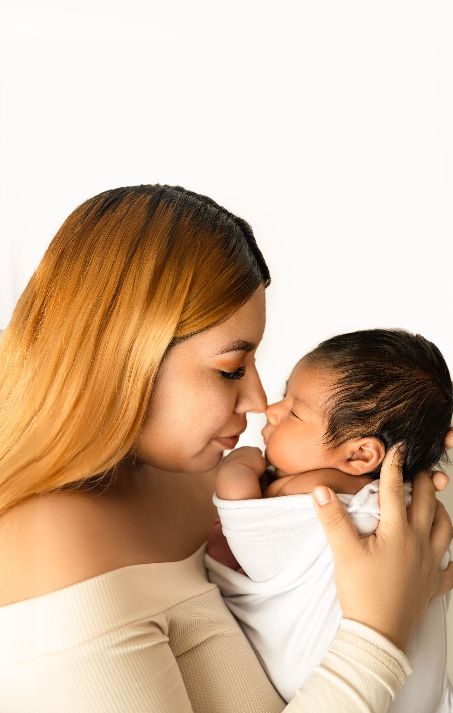 A young mother tenderly touches noses with her swaddled newborn baby against a white background.