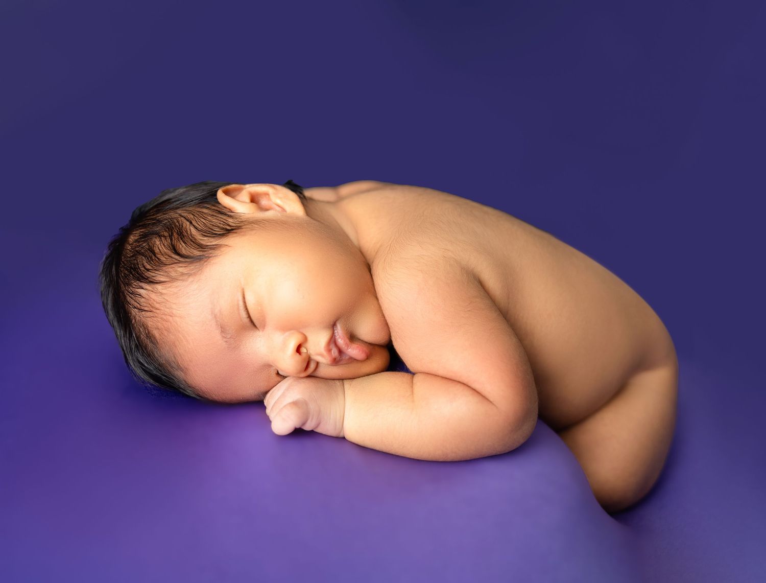 Sleeping newborn baby curled up on purple background in professional studio photo.
