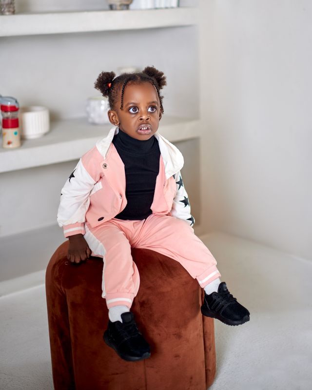 Toddler girl in pink star bomber jacket and matching pants sitting on brown velvet ottoman.