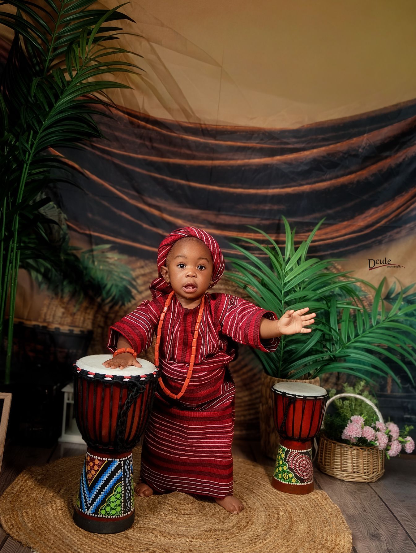 An adorable baby in a red African traditional family photoshoot outfit in Toronto sits between two djembe drums in studio setting.