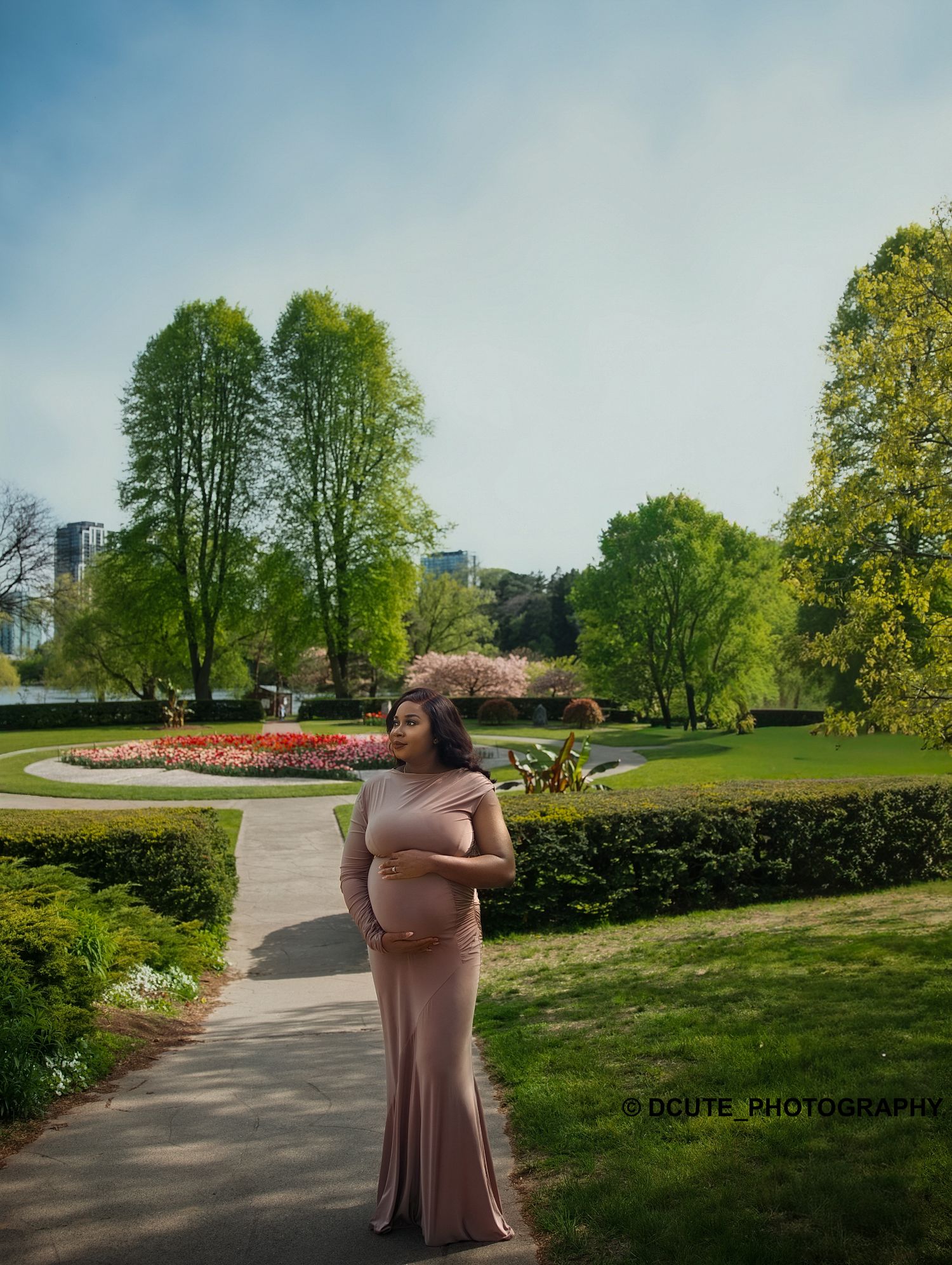 Pregnant woman in elegant beige gown standing on garden path surrounded by colorful flowers during maternity gown photoshoot toronto