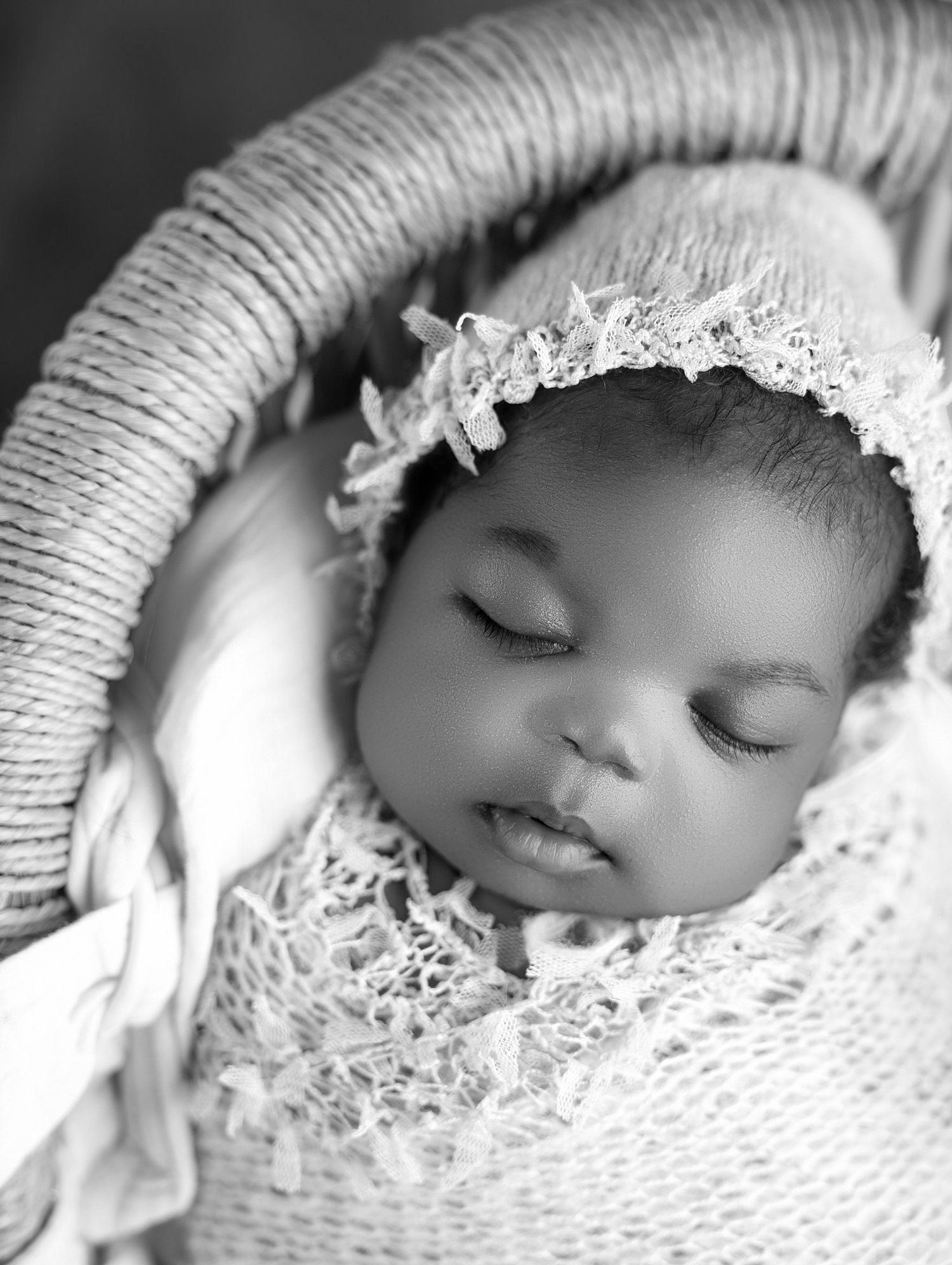 Peaceful sleeping newborn baby wrapped in lace swaddle with floral bonnet in black and white photo.
