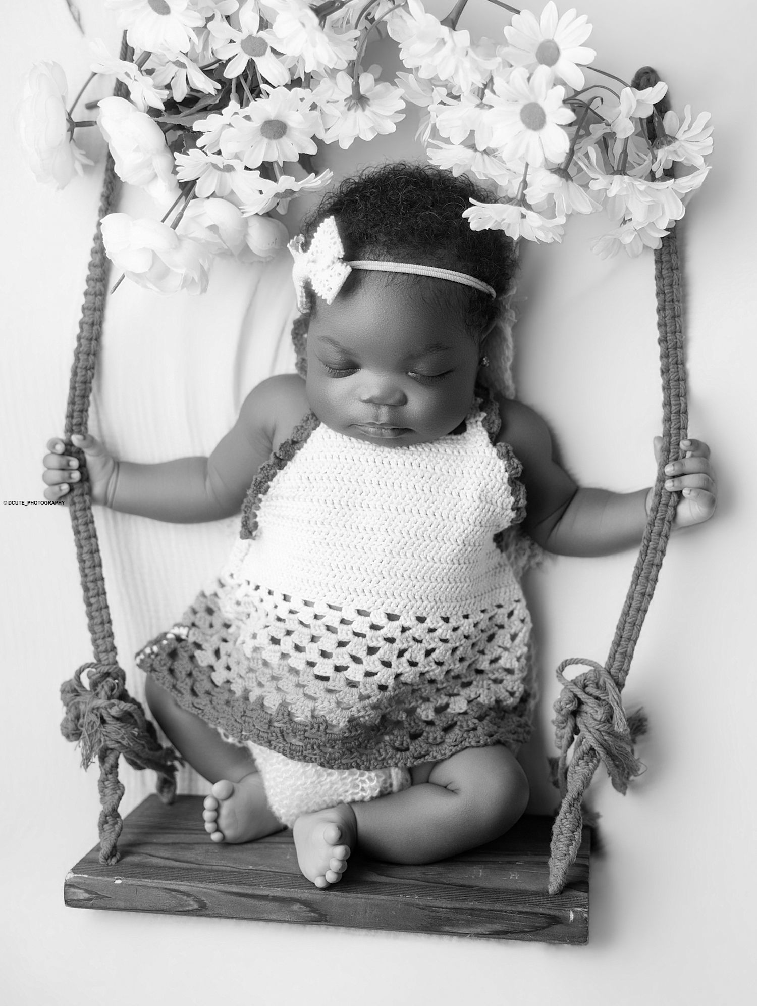 Sleeping newborn baby girl in crochet dress on wooden swing with flowers, black and white portrait photo.
