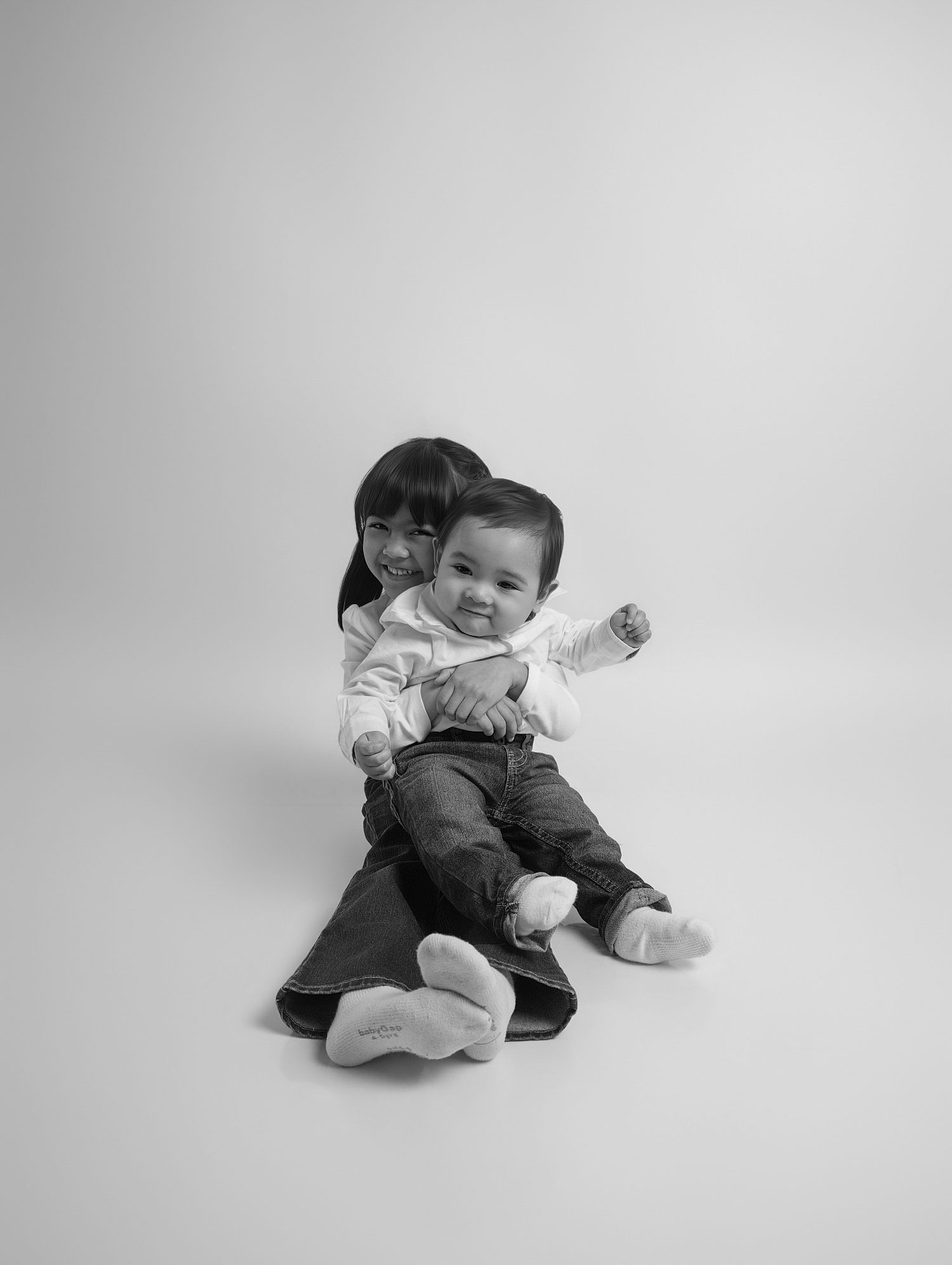 Black and white portrait of young girl hugging smiling toddler brother on floor.
