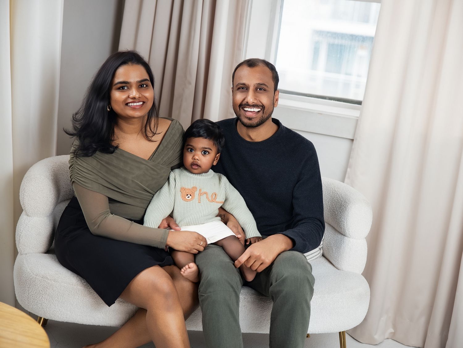 Happy South Asian family of three sitting together on a white chair in a bright, modern indoor setting in Downtown Toronto family photograph