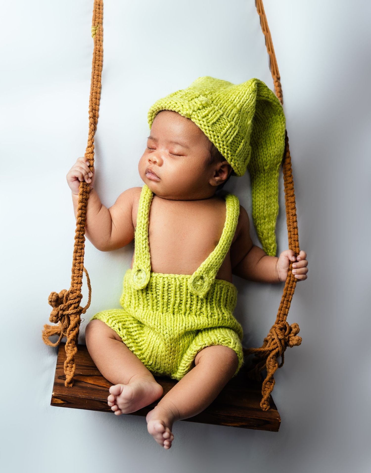 Sleeping newborn baby wearing green knit elf hat and romper outfit, resting on a wooden swing with rope.