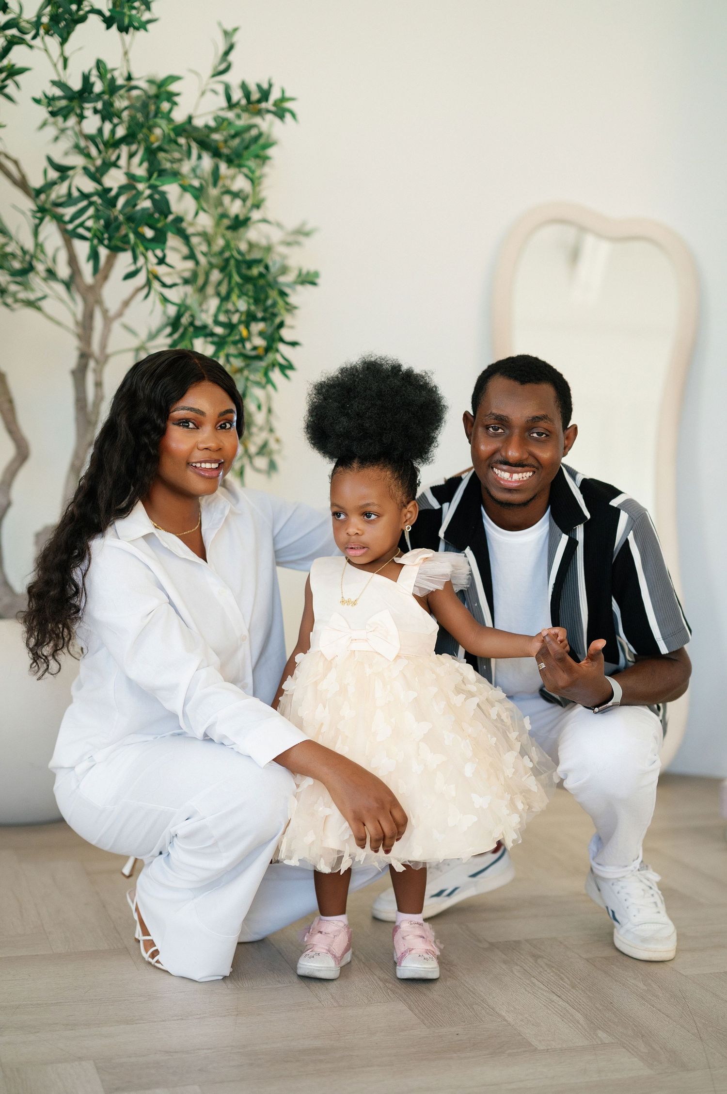 Mother, father, and toddler daughter in white outfits pose indoors. The lady is a professional children's photographer in Toronto